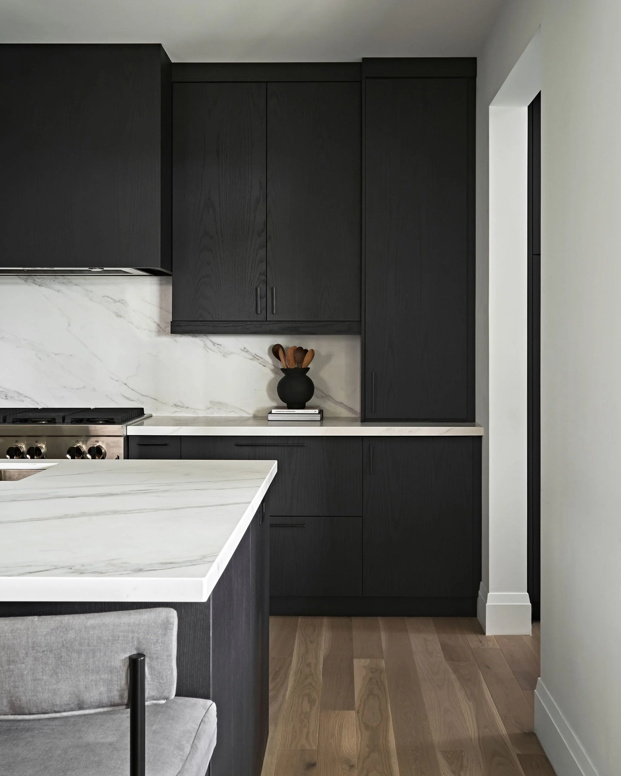 Modern kitchen with black cabinets, marble countertop, and wooden flooring. A black vase with wooden utensils is on the counter.