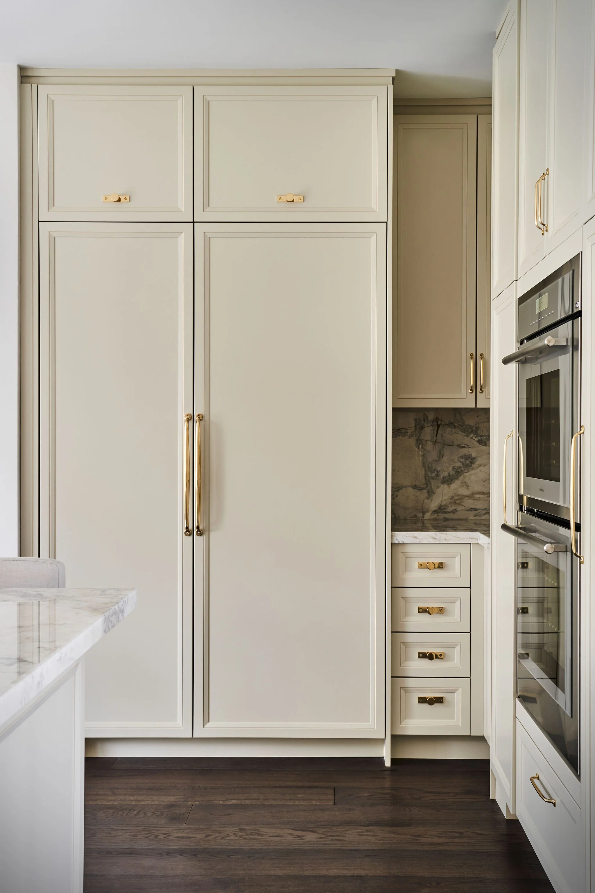 A kitchen with cream-colored cabinets and a marble countertop. There are gold handles on the cabinets and drawers, with dark wood flooring.
