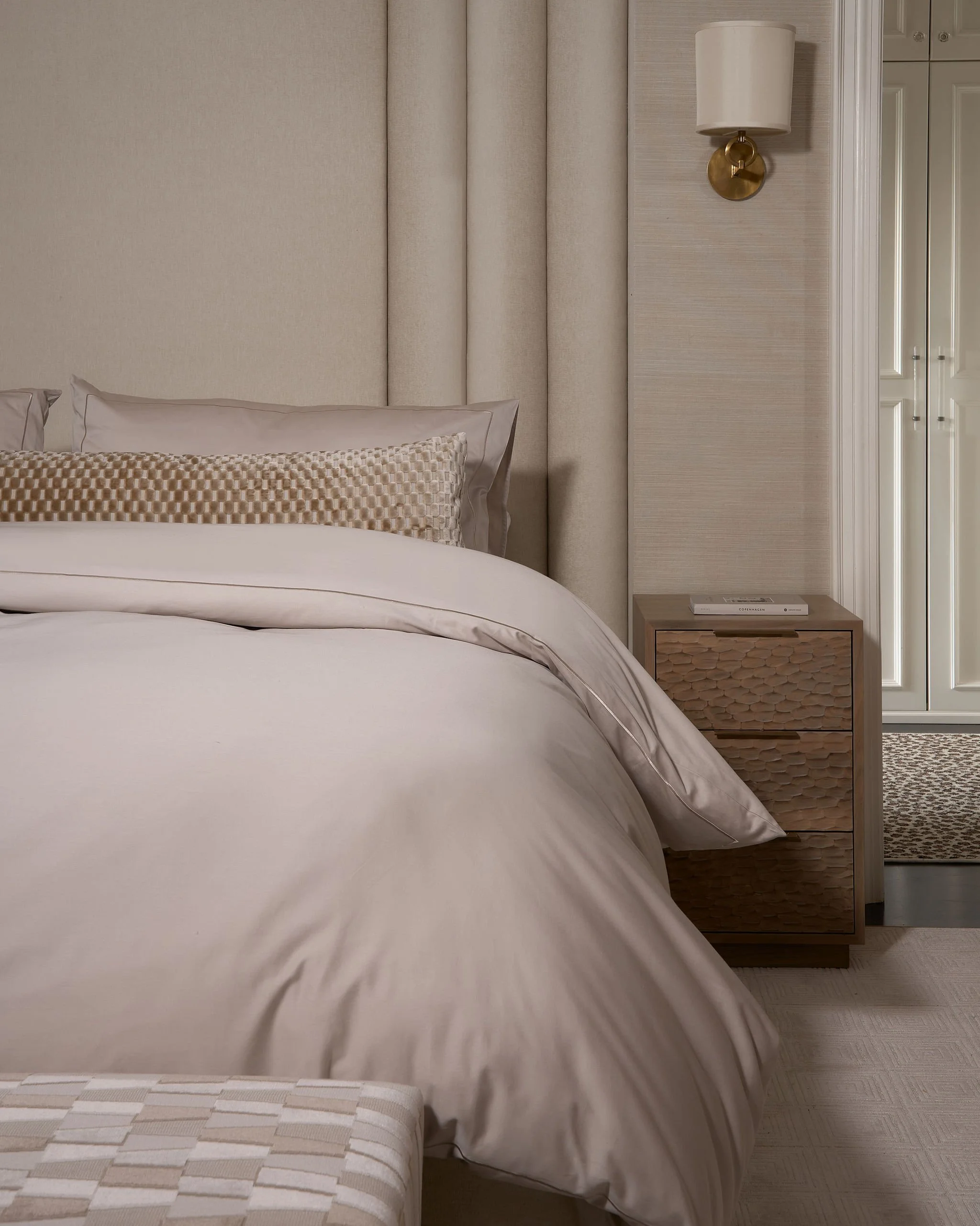 A bedroom with a bed, beige headboard, and a beige duvet. There are two pillows, one with a textured pattern and the other with plain fabric. A nightstand with a book on it is beside the bed, and a wall-mounted light with a white lampshade and a bras