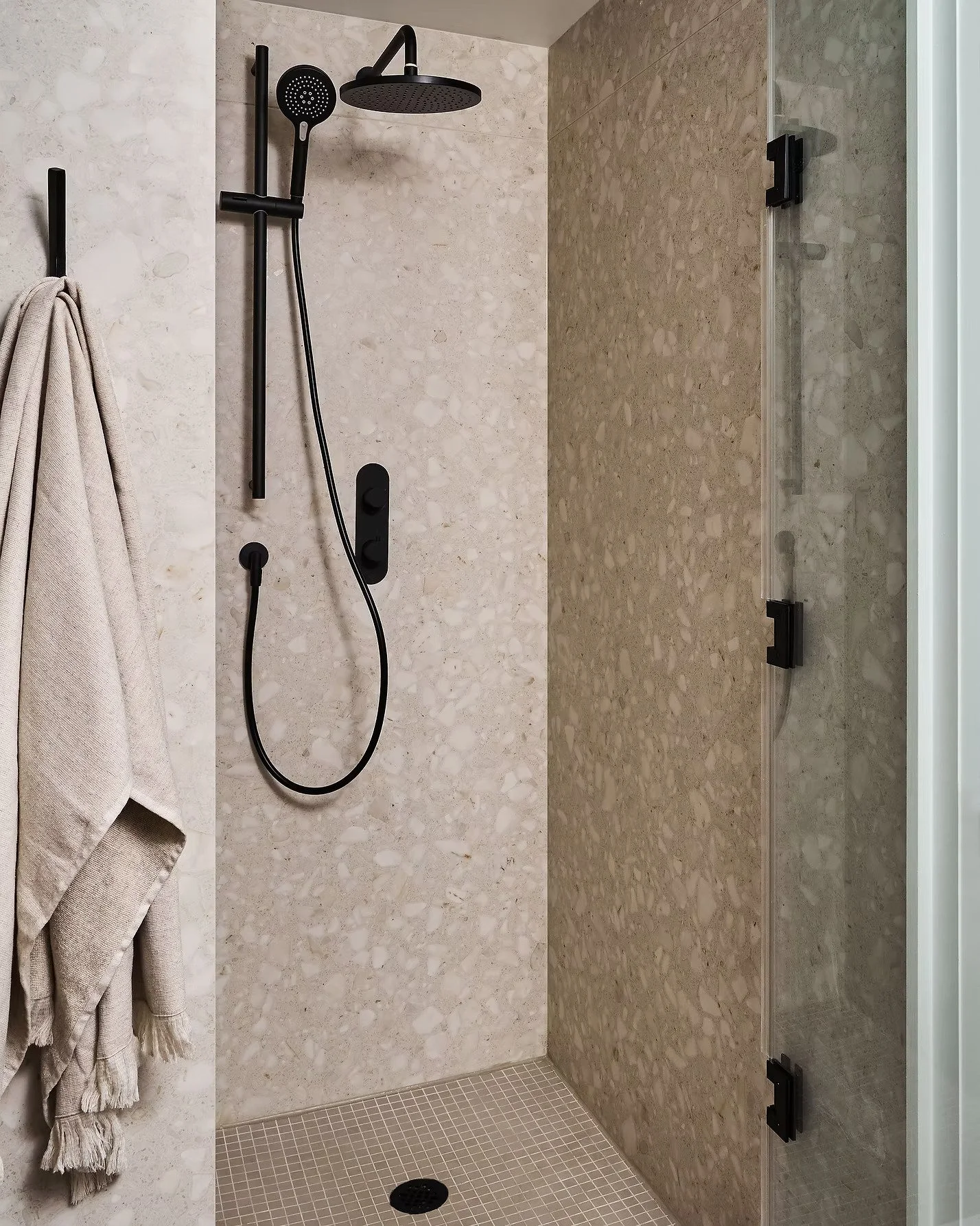 A modern shower area with beige and brown stone tiles, black shower fixtures, a rainfall showerhead, a handheld shower, a wall-mounted control panel, a glass shower door with black hinges, and beige towels hanging on a hook.