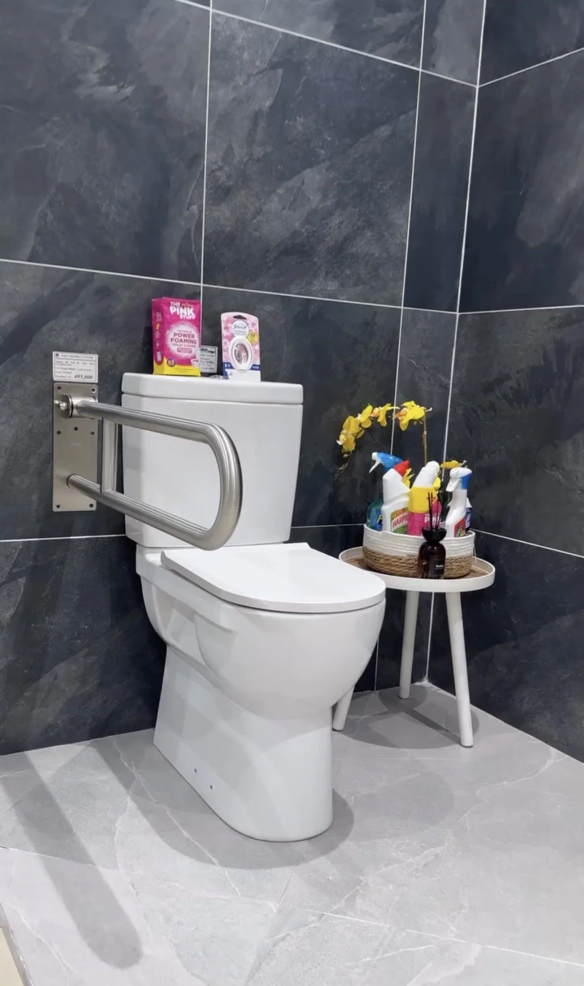 Bathroom with dark gray tiled walls, a white toilet with a safety rail, and a small white table holding cleaning supplies and yellow flowers in a black vase.
