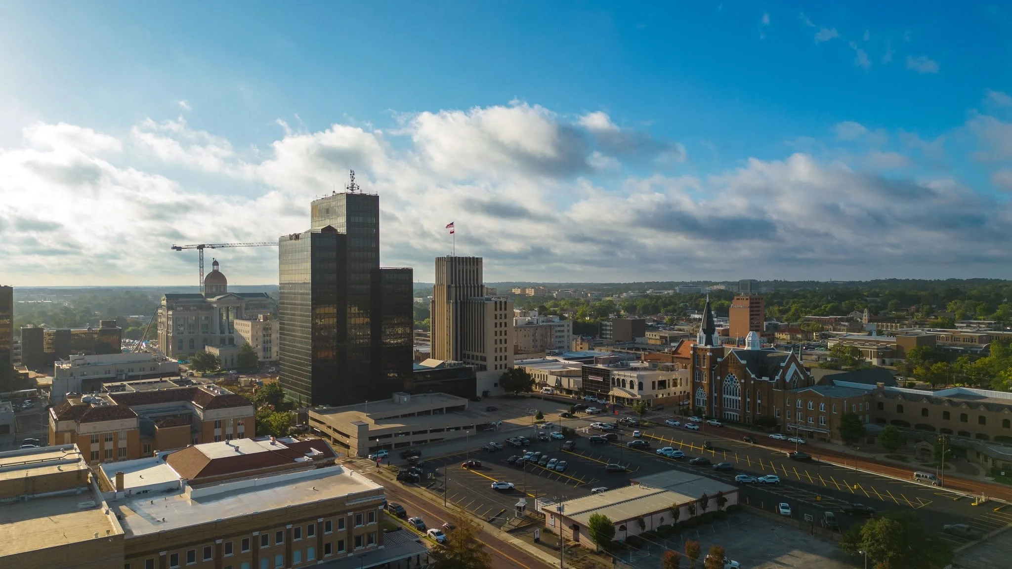 City skyline with tall modern buildings, a construction crane, historic buildings, parking lots, and a green landscape in the background under a partly cloudy sky.