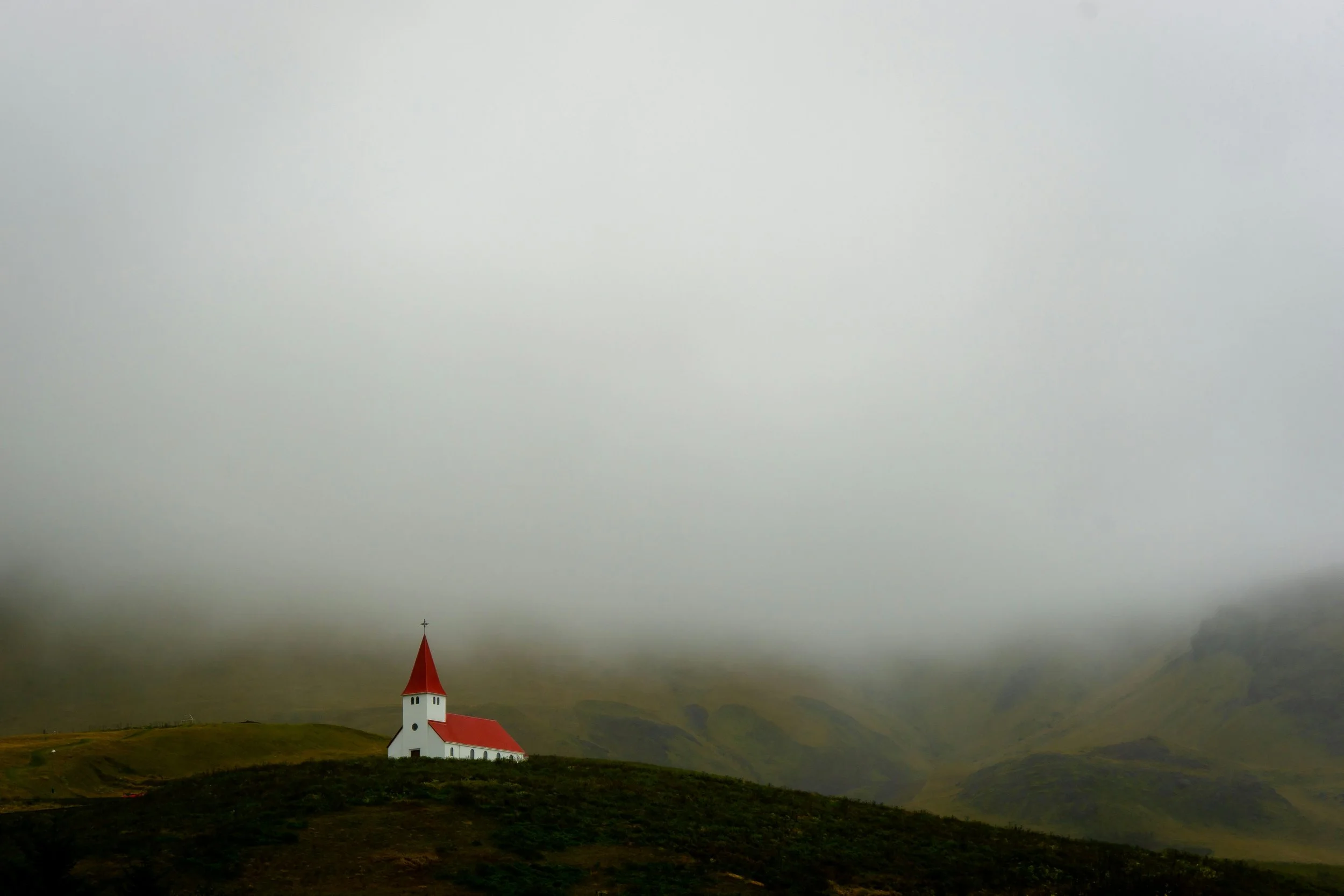 A small white church with a red roof and steeple is situated on a green hill under an overcast sky, with faint distant hills in the background.