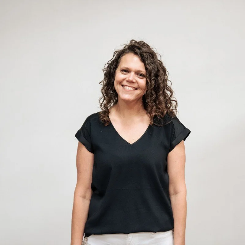 A woman with curly brown hair wearing a black V-neck T-shirt and light-colored pants, smiling at the camera against a plain white background.