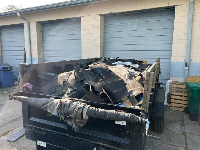 Overflowing dumpster filled with crumpled cardboard and black plastic debris parked in front of a building with closed garage doors.