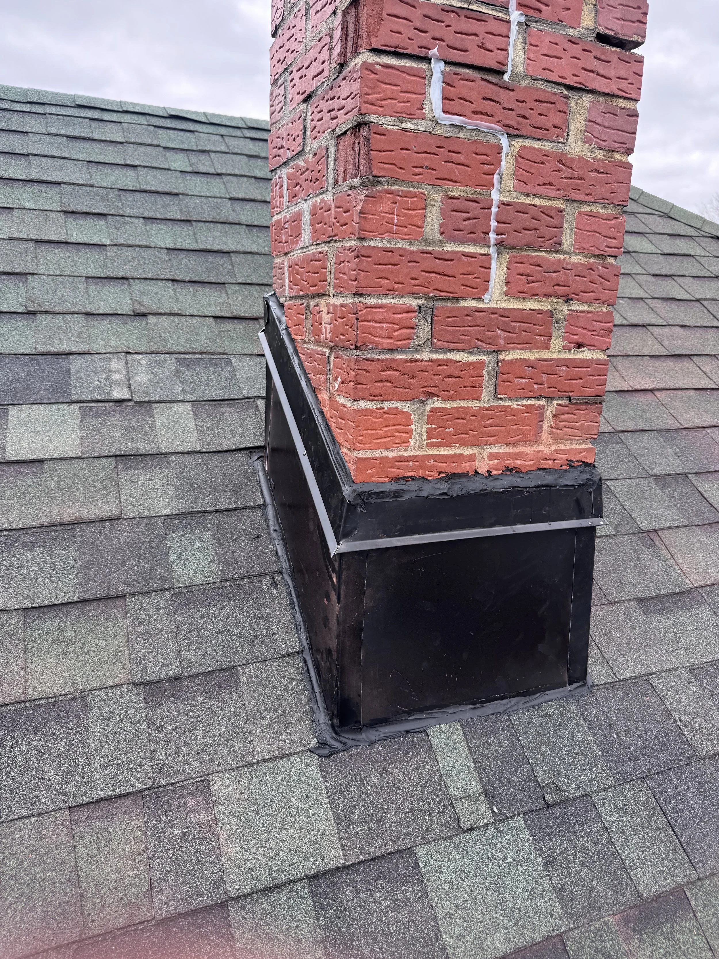 Close-up of a brick chimney on a roof with dark shingles, with black flashing and sealant at the base.