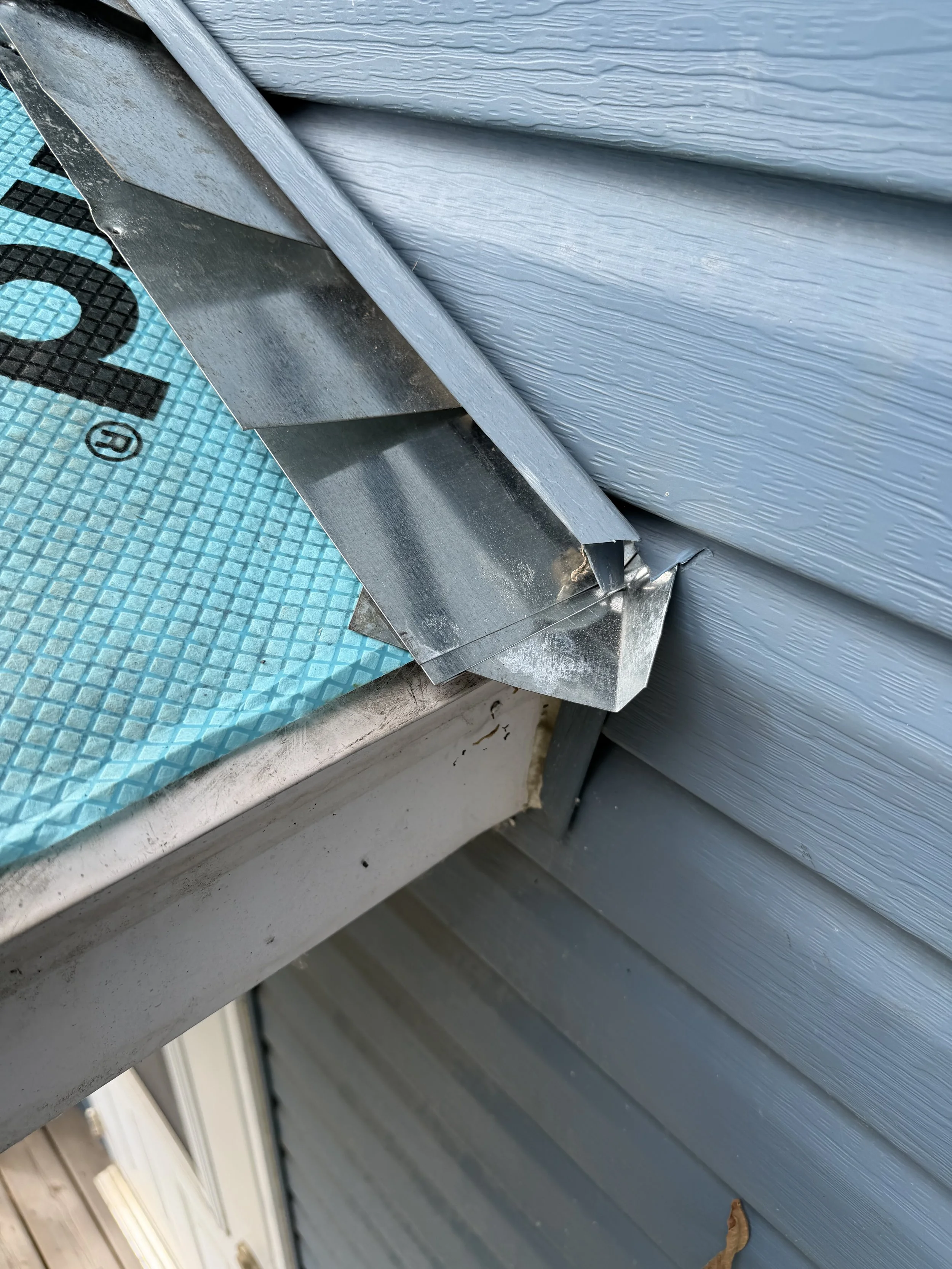 Close-up of the corner of a house with vinyl siding, a metal flashing, and part of a doormat with a textured blue surface and a black number 2.