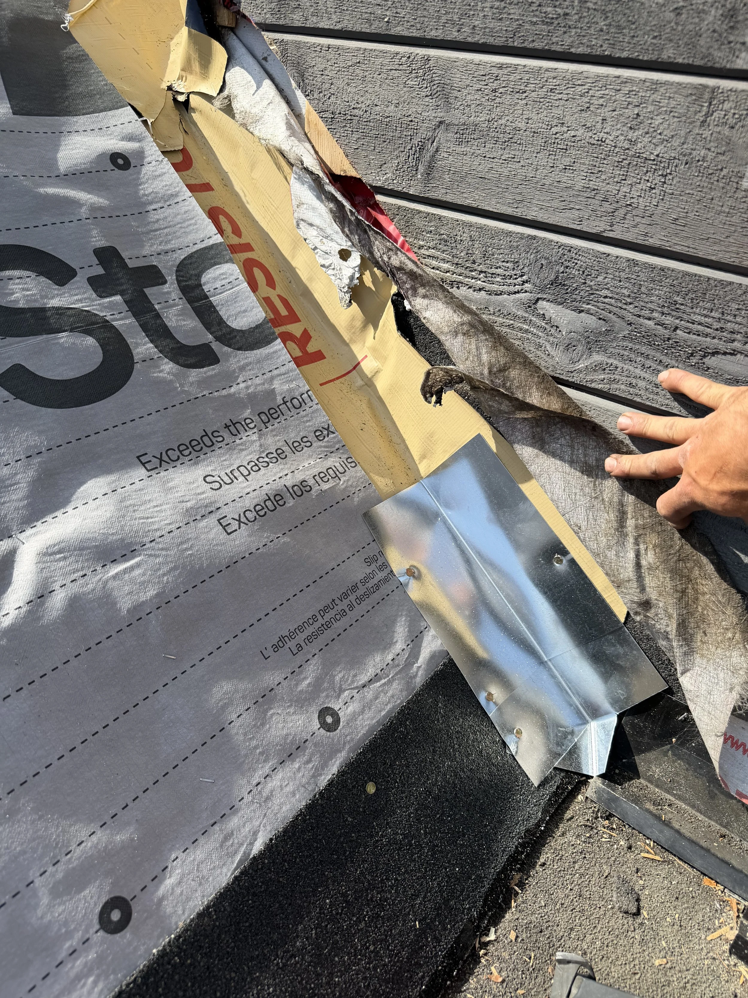 Close-up of a construction site showing a damaged section of house siding and the flashing beneath it, with a person's hand pointing at the damage.