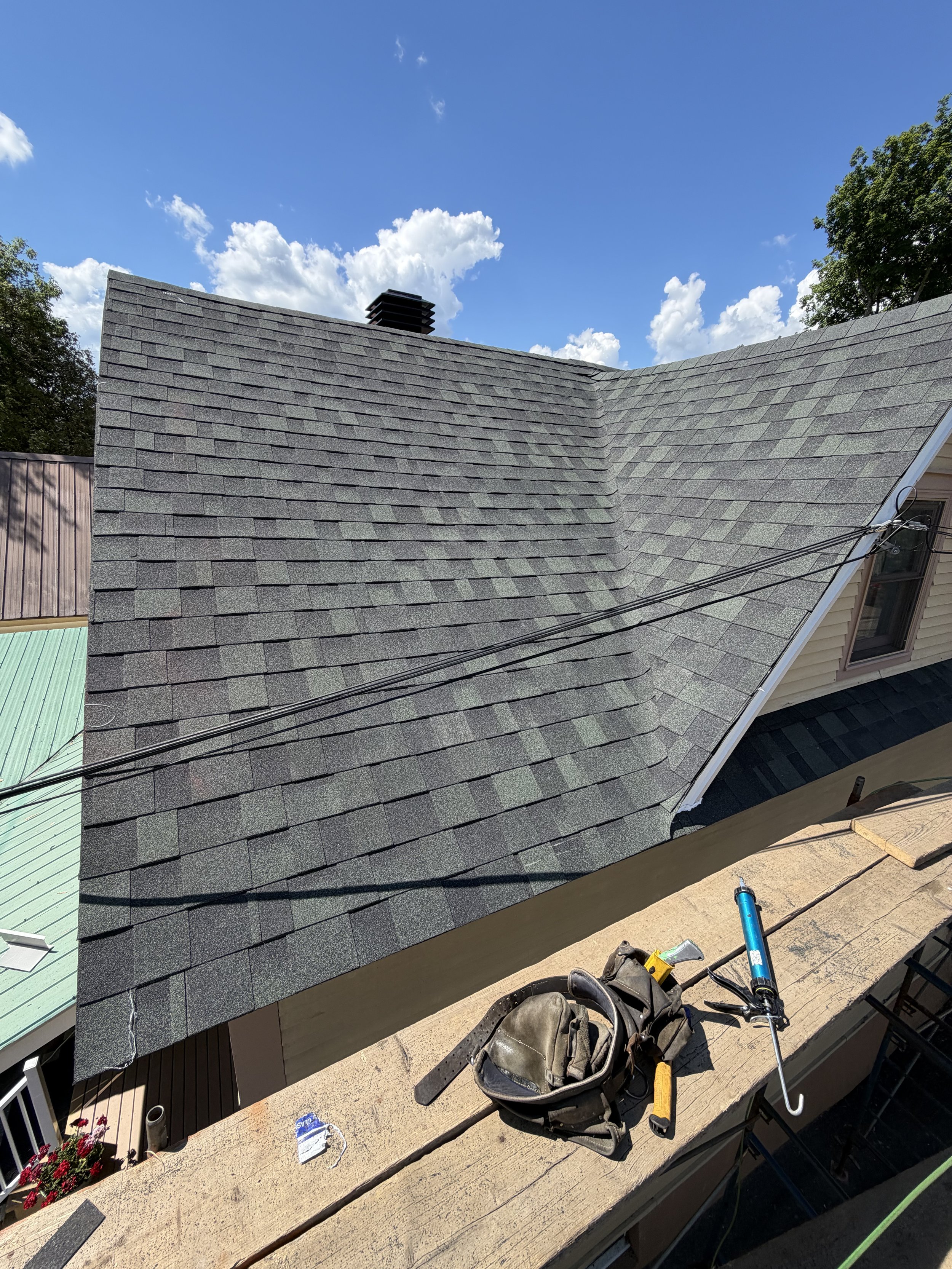 View of a house roof with gray shingles, a chimney, and some tools and equipment on the balcony, with clear skies and some trees in the background.