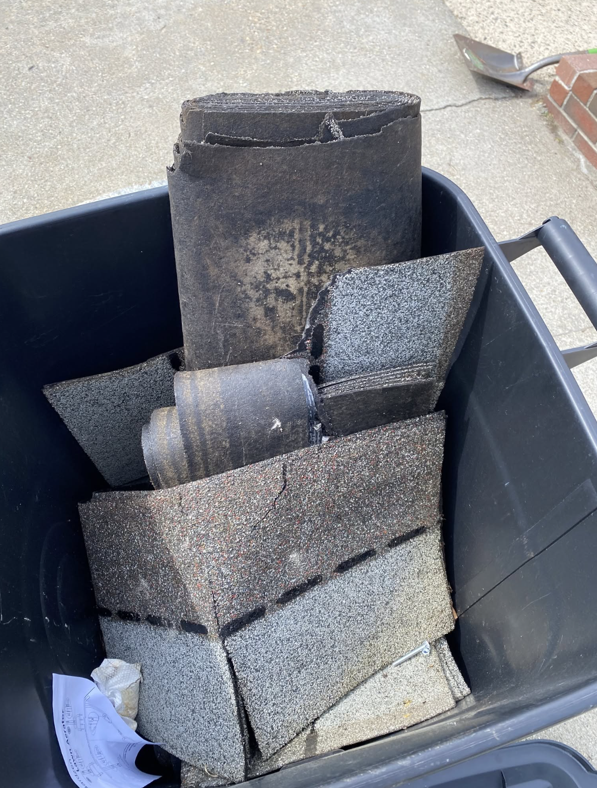 A black trash bin filled with debris including a large charred roll of roofing felt and broken concrete pavers. A shovel is on the ground nearby, and there is some paper or tissue in the trash.