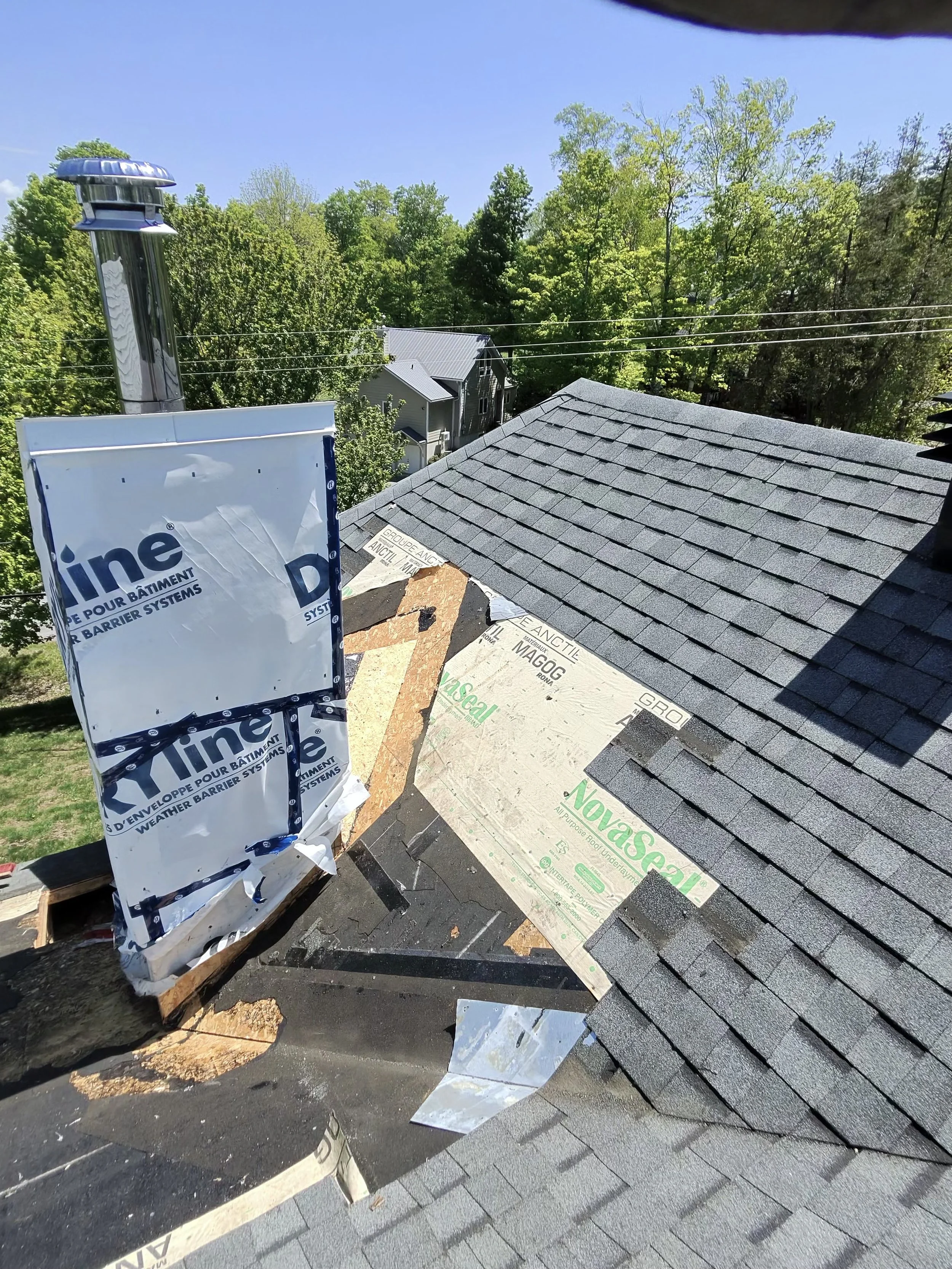 View of a residential rooftop under construction with roofing materials and insulation, surrounded by green trees and a clear blue sky.