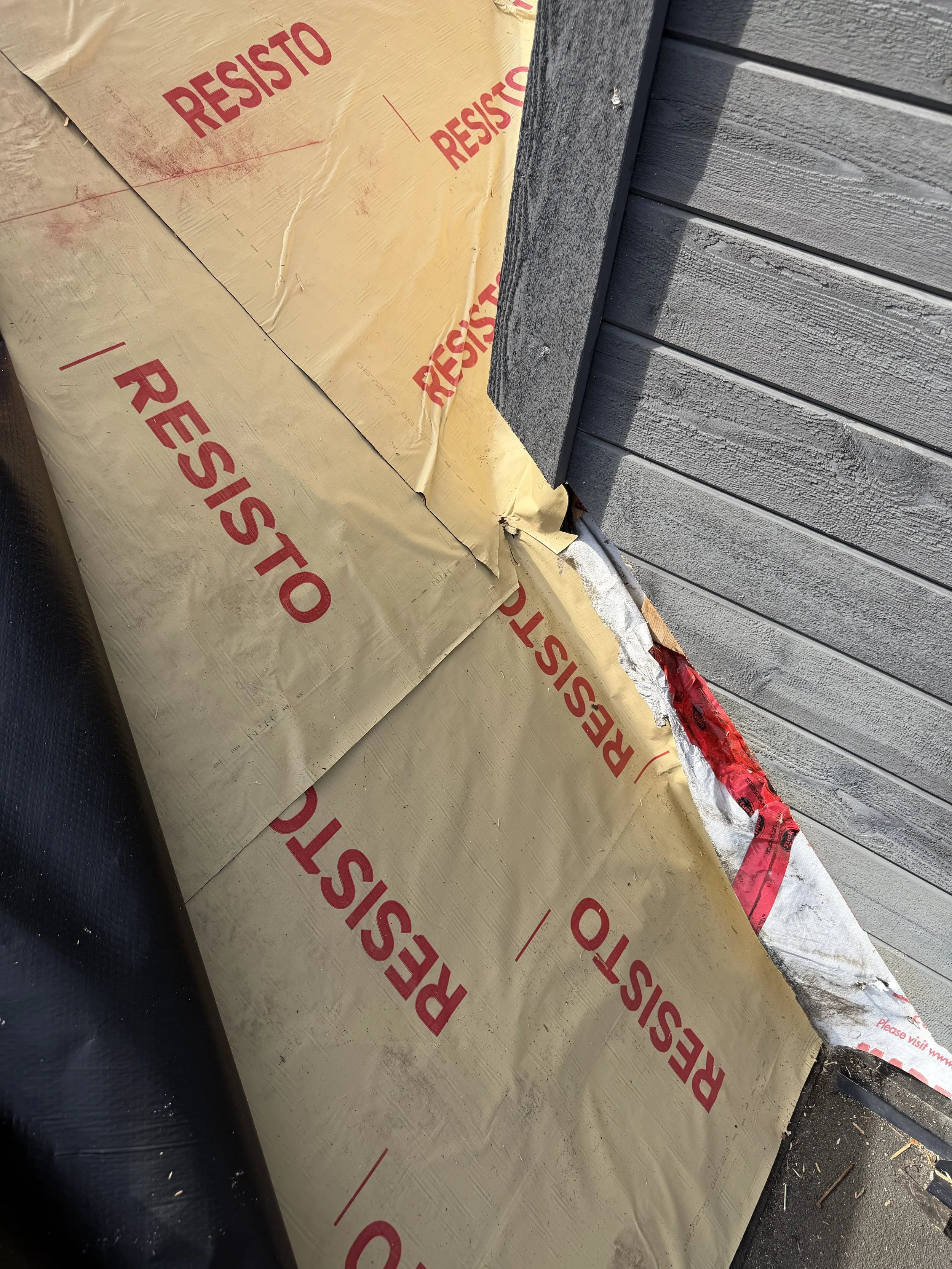 Close-up of house siding with insulation material partially installed next to a black pipe and a red and white strip of protective tape on the ground.