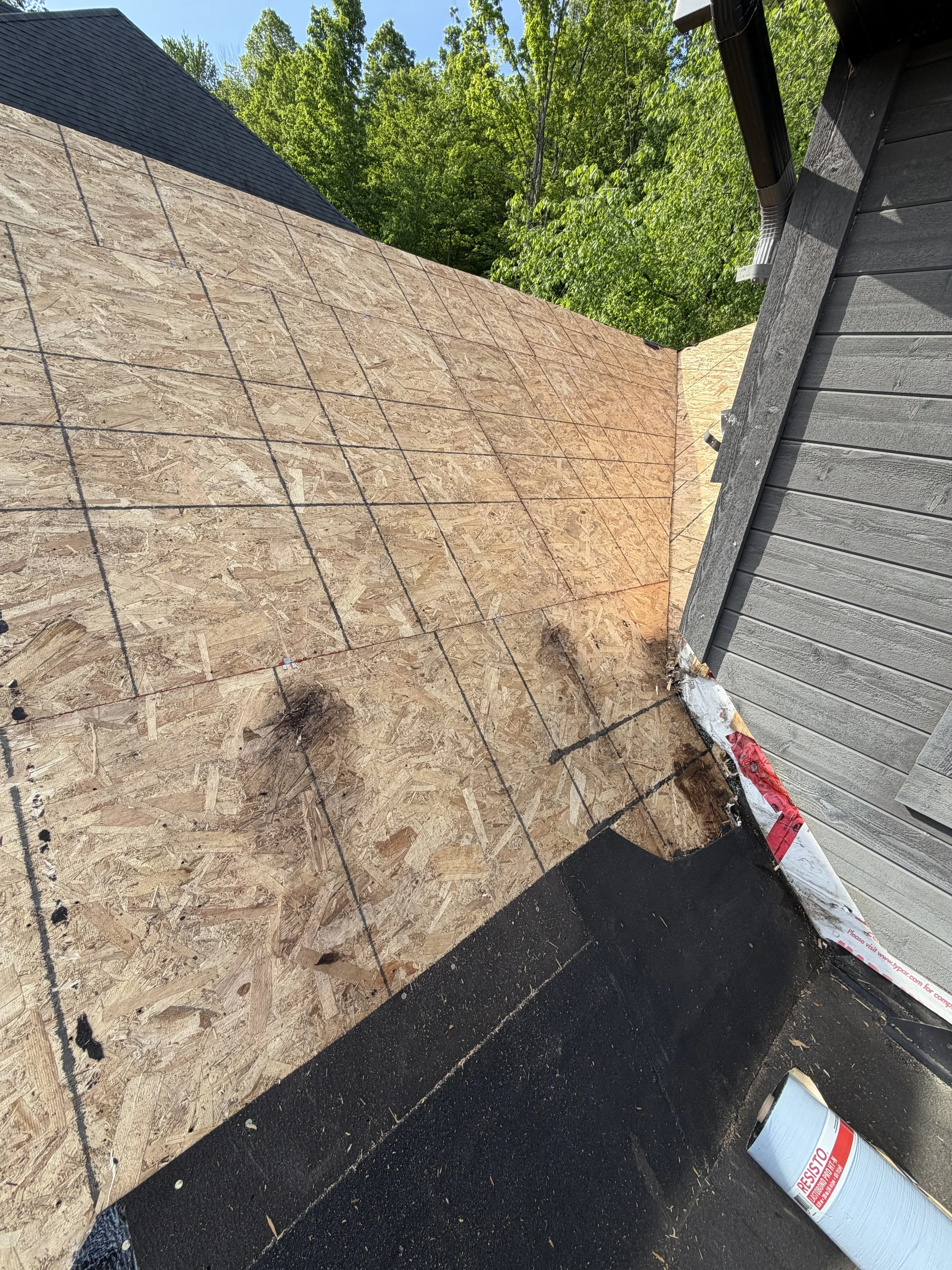 The image shows a section of a house roof where the shingles have been removed, exposing oriented strand board (OSB) sheathing. There is construction material and a can of required spray or sealant on the black surface below, with green trees in the background.