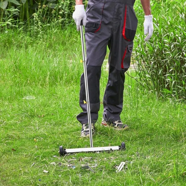 Person wearing work gloves using a metal detector to scan the grass in a grassy outdoor area surrounded by tall plants.
