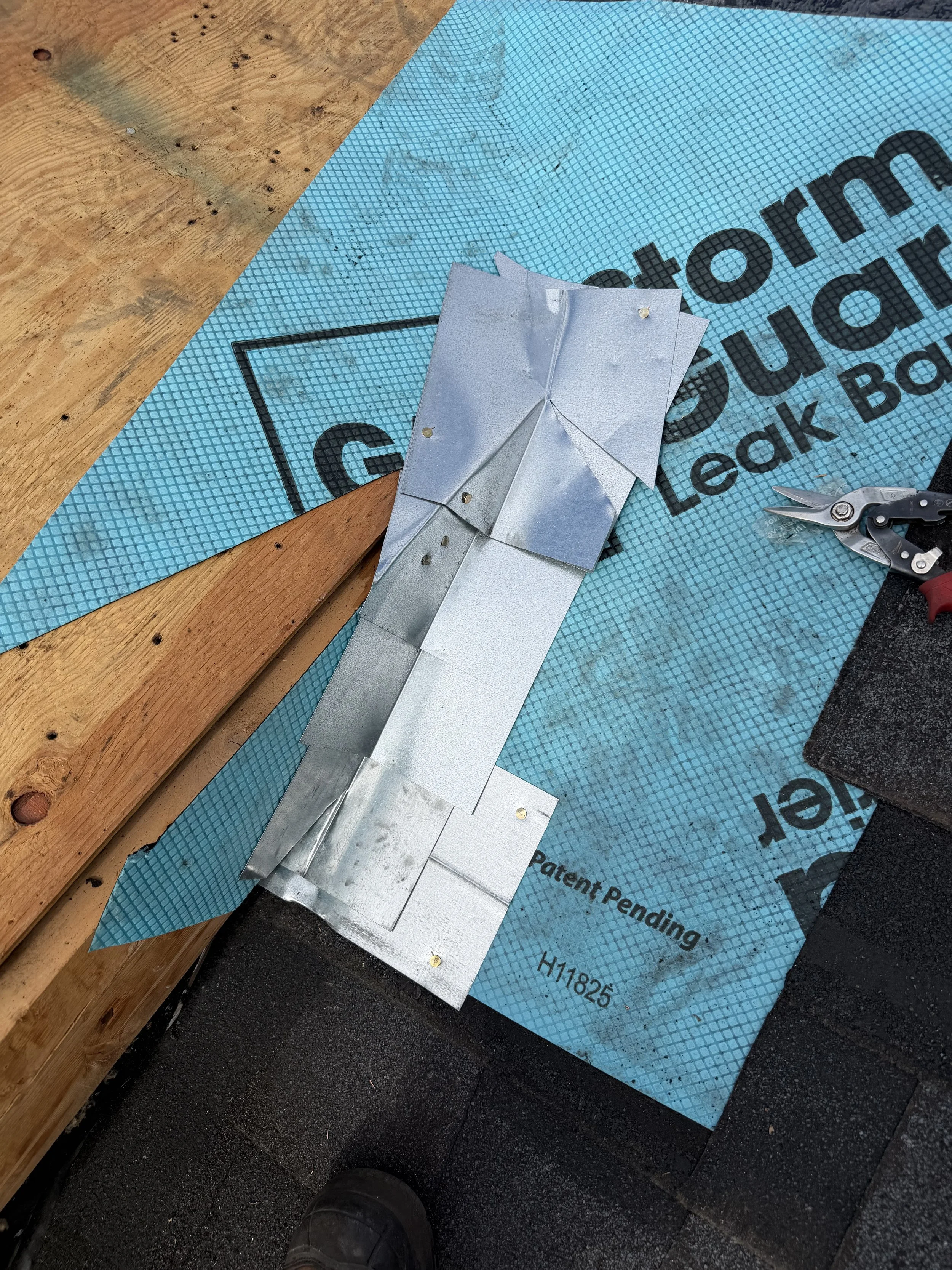 Metal flashing or roofing material laid over blue roofing underlayment on a construction site, with a wooden surface to the left and a black asphalt surface below, a pair of pliers or snips on the right.
