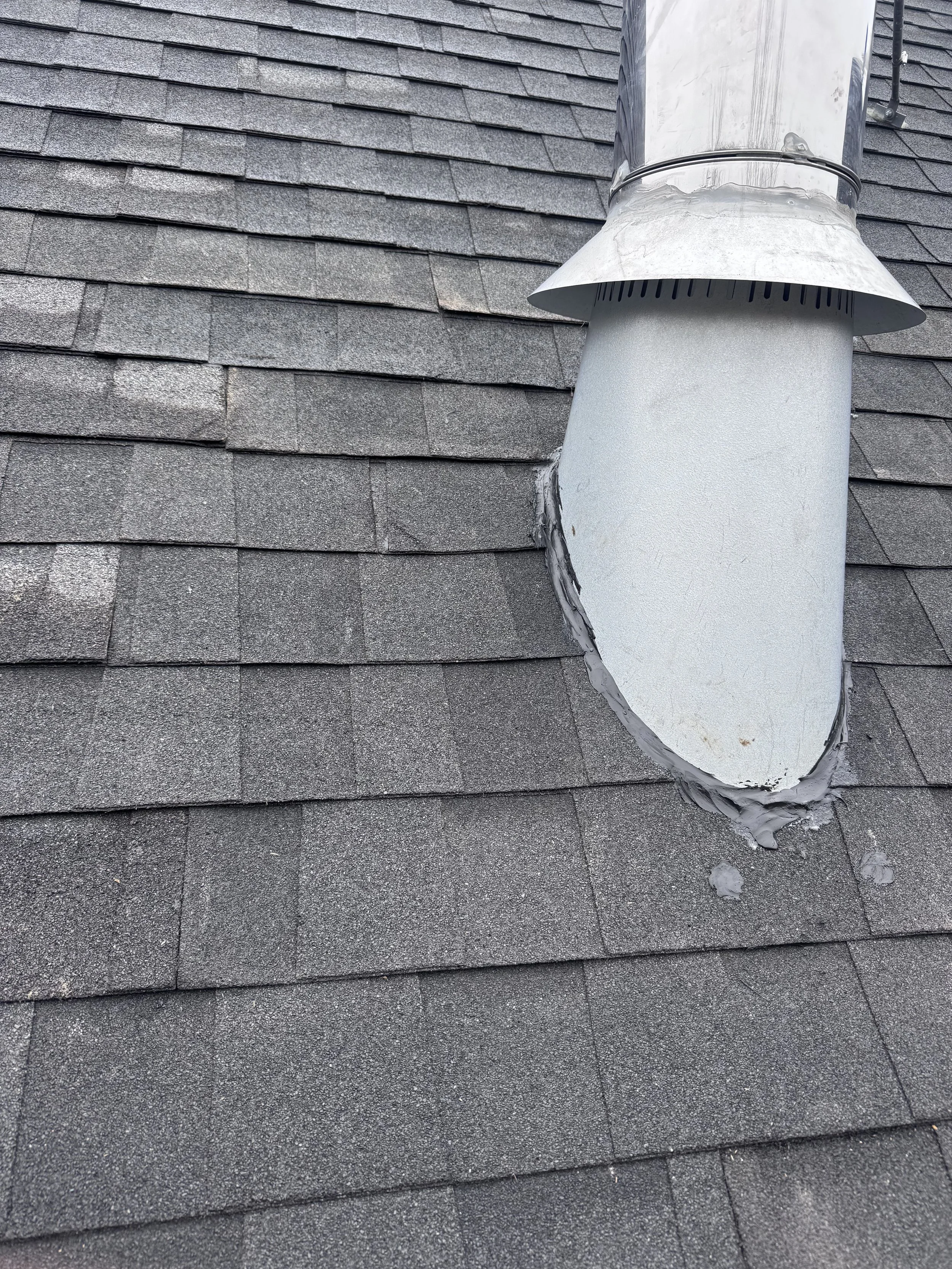 A roof vent pipe protruding from a gray shingled roof, covered with a metal cap and flashing to seal around the pipe.