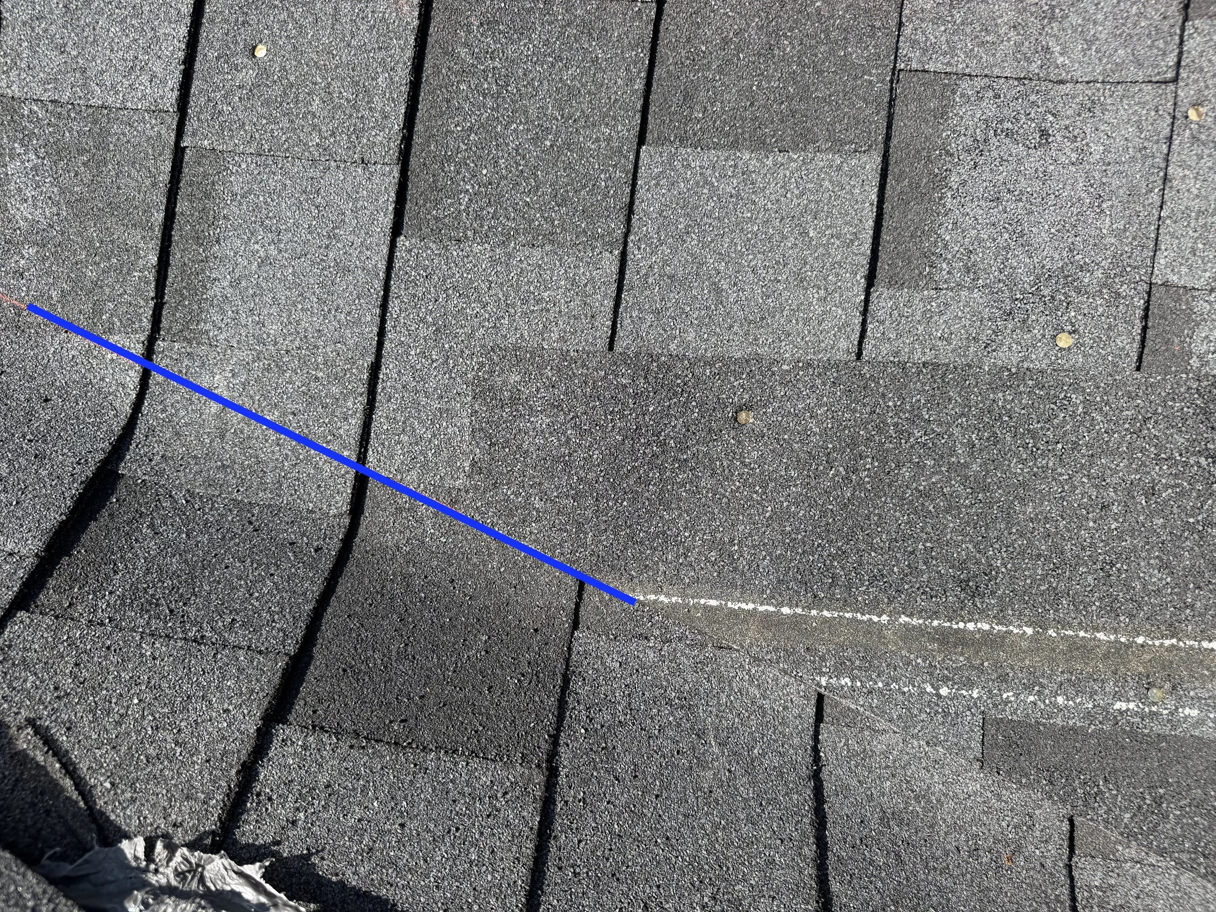 Close-up of black asphalt shingles with a blue chalk line across them, and a small piece of crumpled paper at the bottom left corner.