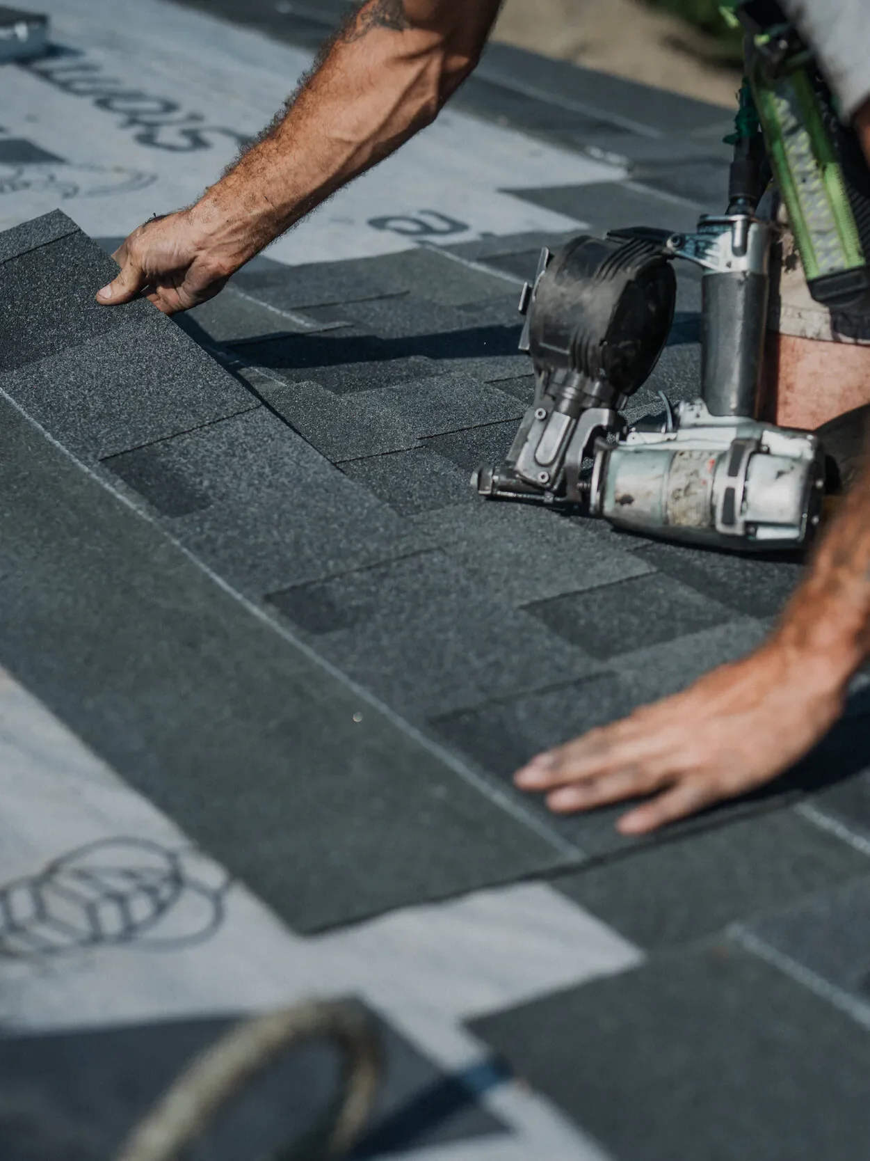 A person installing asphalt shingles on a roof, using a nail gun and wearing work gloves.