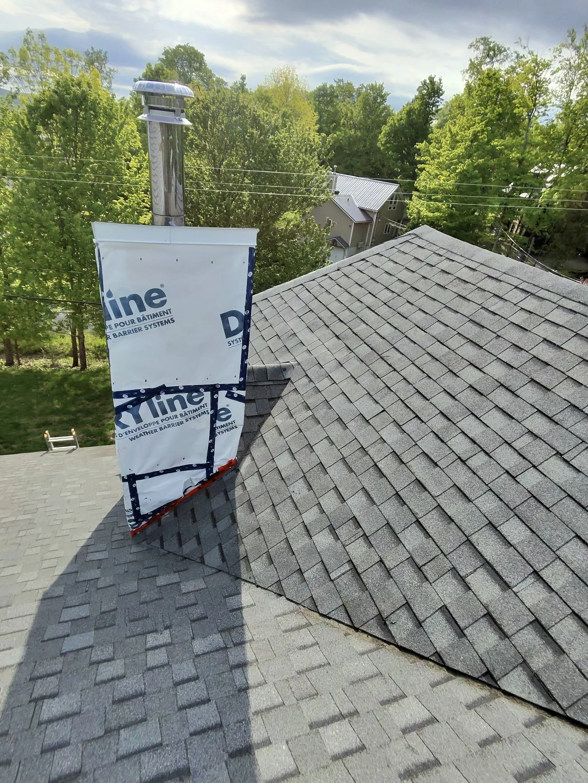Shingled roof of a house with weather barrier system and vent pipe. Trees and other houses are visible in the background.