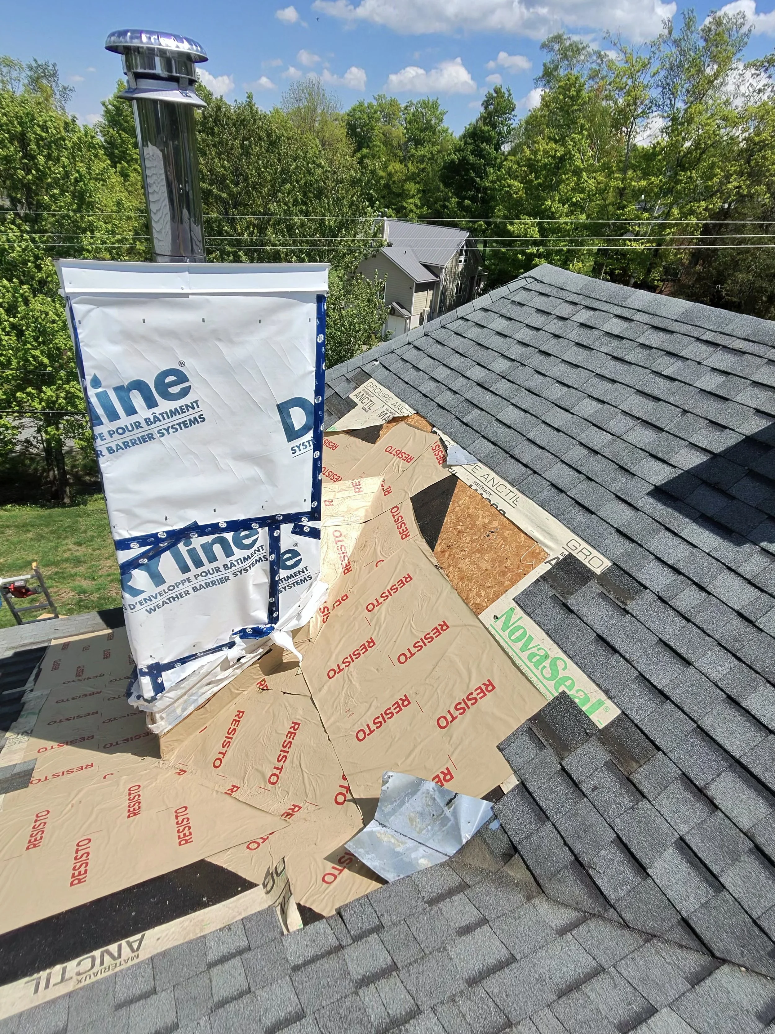 View from a rooftop showing a chimney, roofing materials, and shingles, with trees and a cloudy blue sky in the background.