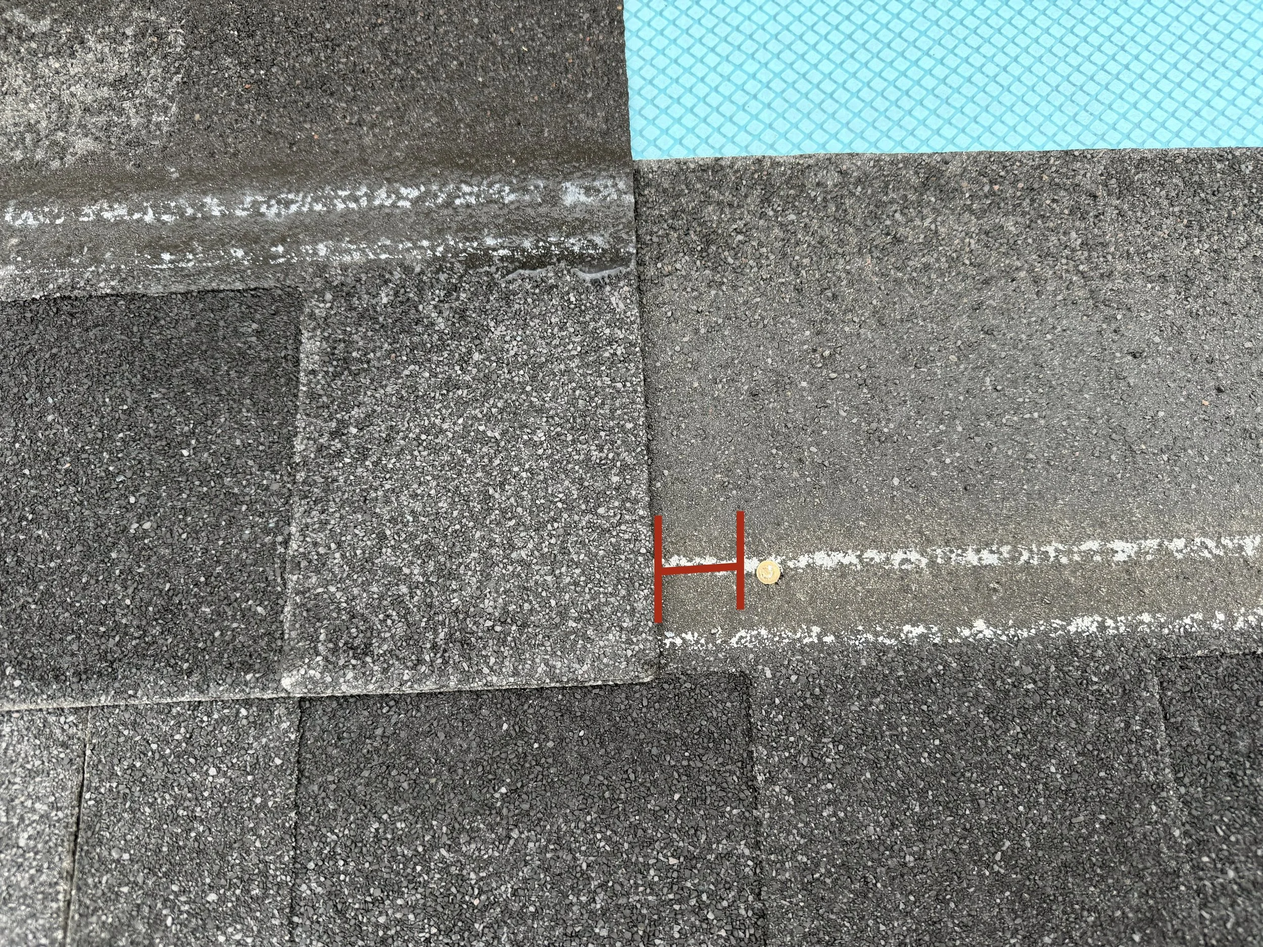 A section of a tennis court with a textured dark gray surface, white boundary lines, and a blue patterned area in the top right corner. A small coin and red markings are visible near the bottom of the image.