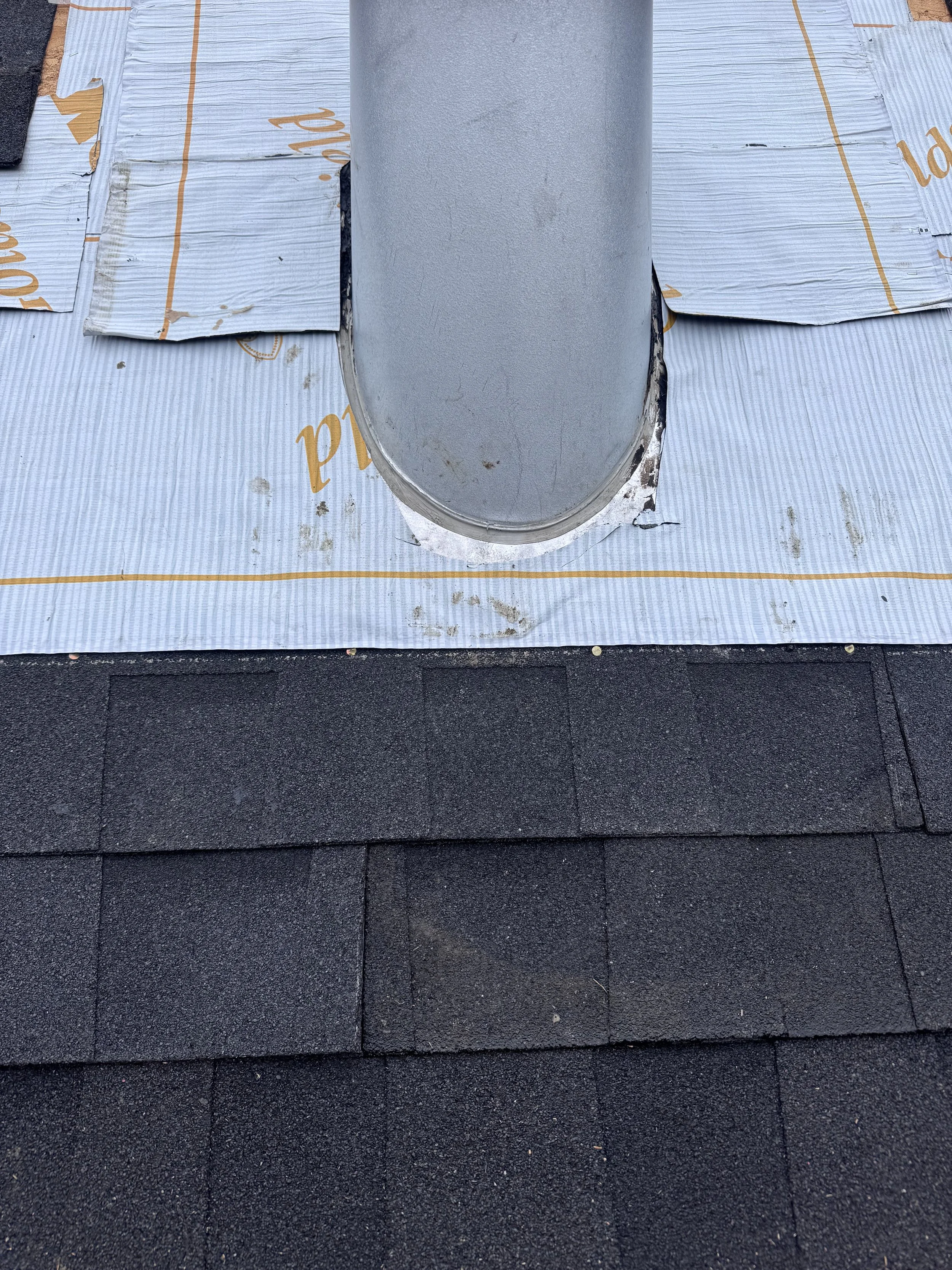 A flat rooftop covered with black asphalt shingles, with a metal vent pipe protruding from the roof and surrounded by a white roofing membrane.