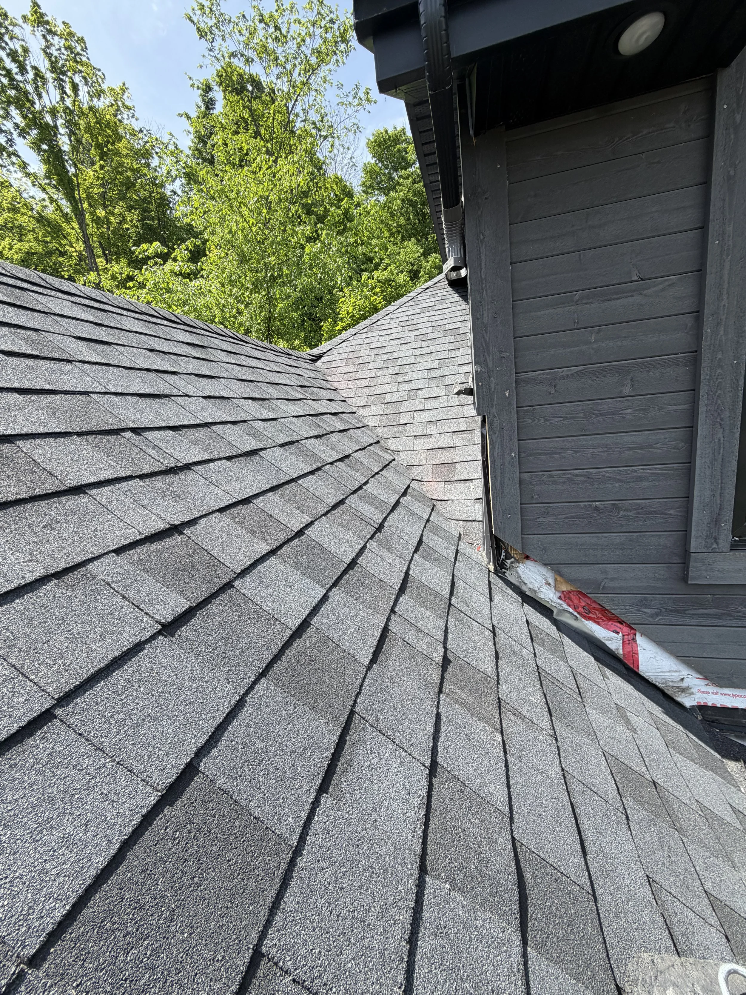 Close-up of a gray shingle roof with a wooden exterior wall and trees in the background under a clear sky.