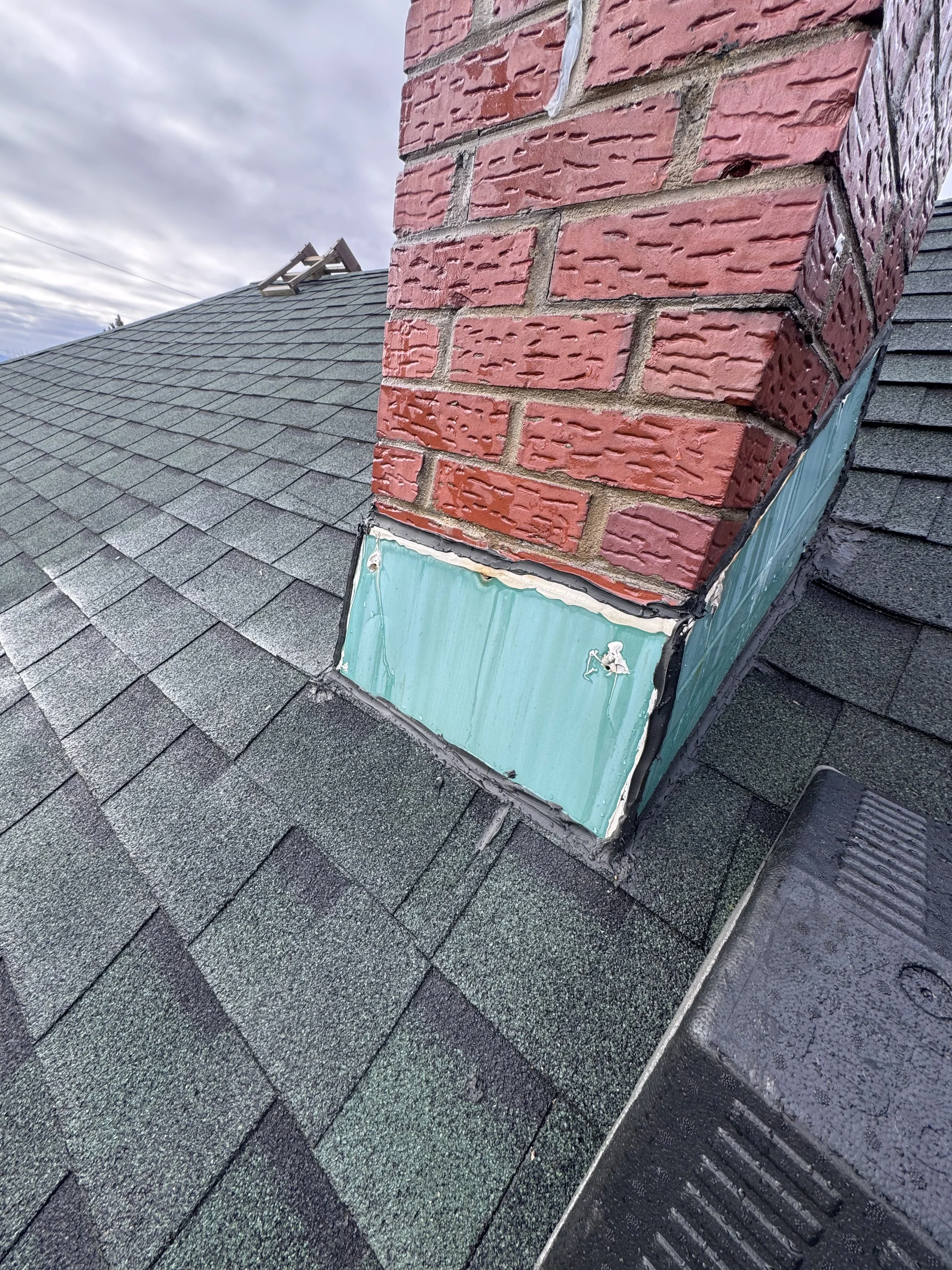 Close-up of a chimney with a brick exterior on a shingled roof, with roofing materials and flashing visible.