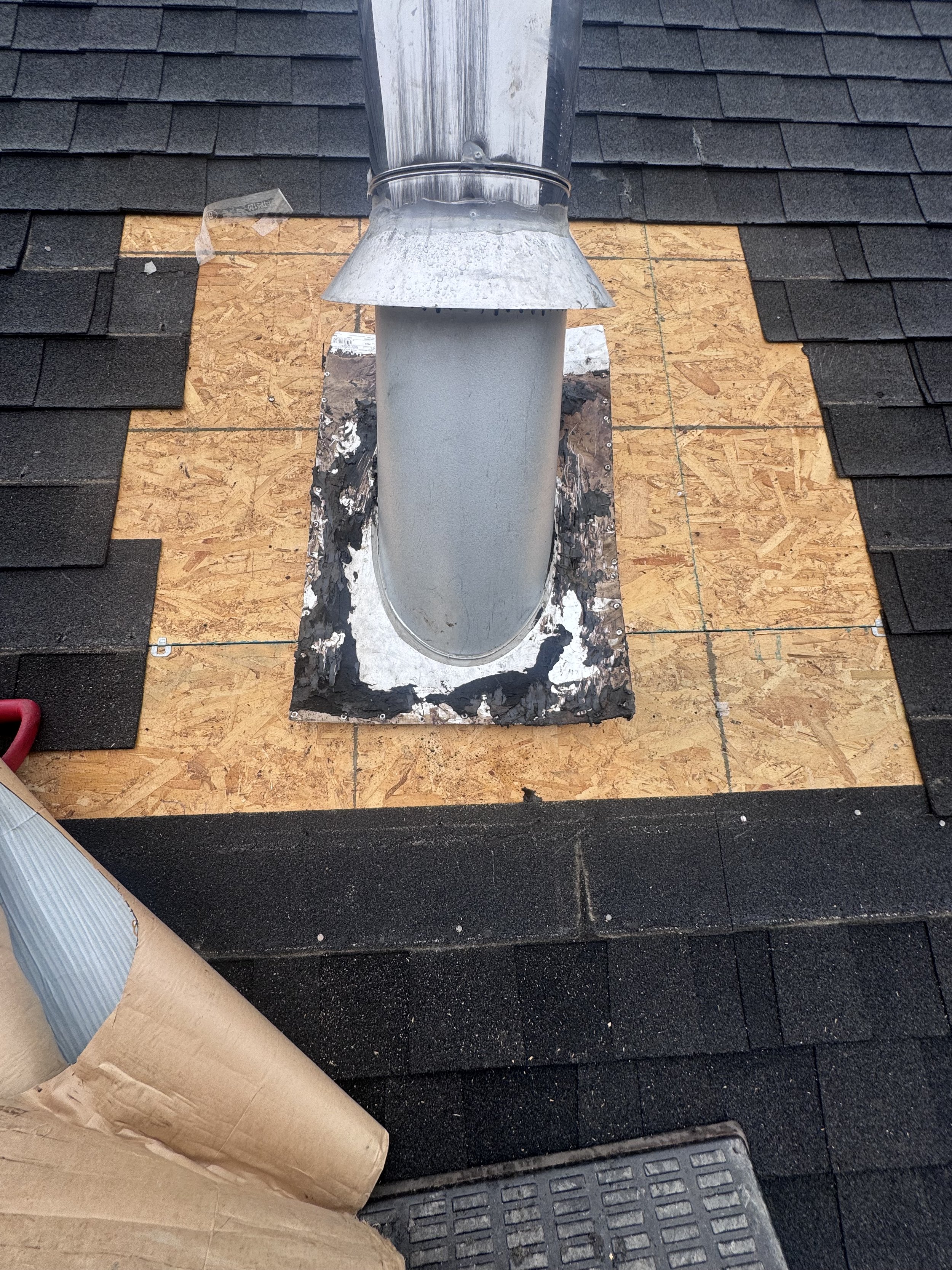 Construction site roof with black shingle tiles, plywood sheathing, and a metal vent pipe, surrounded by tools and materials.