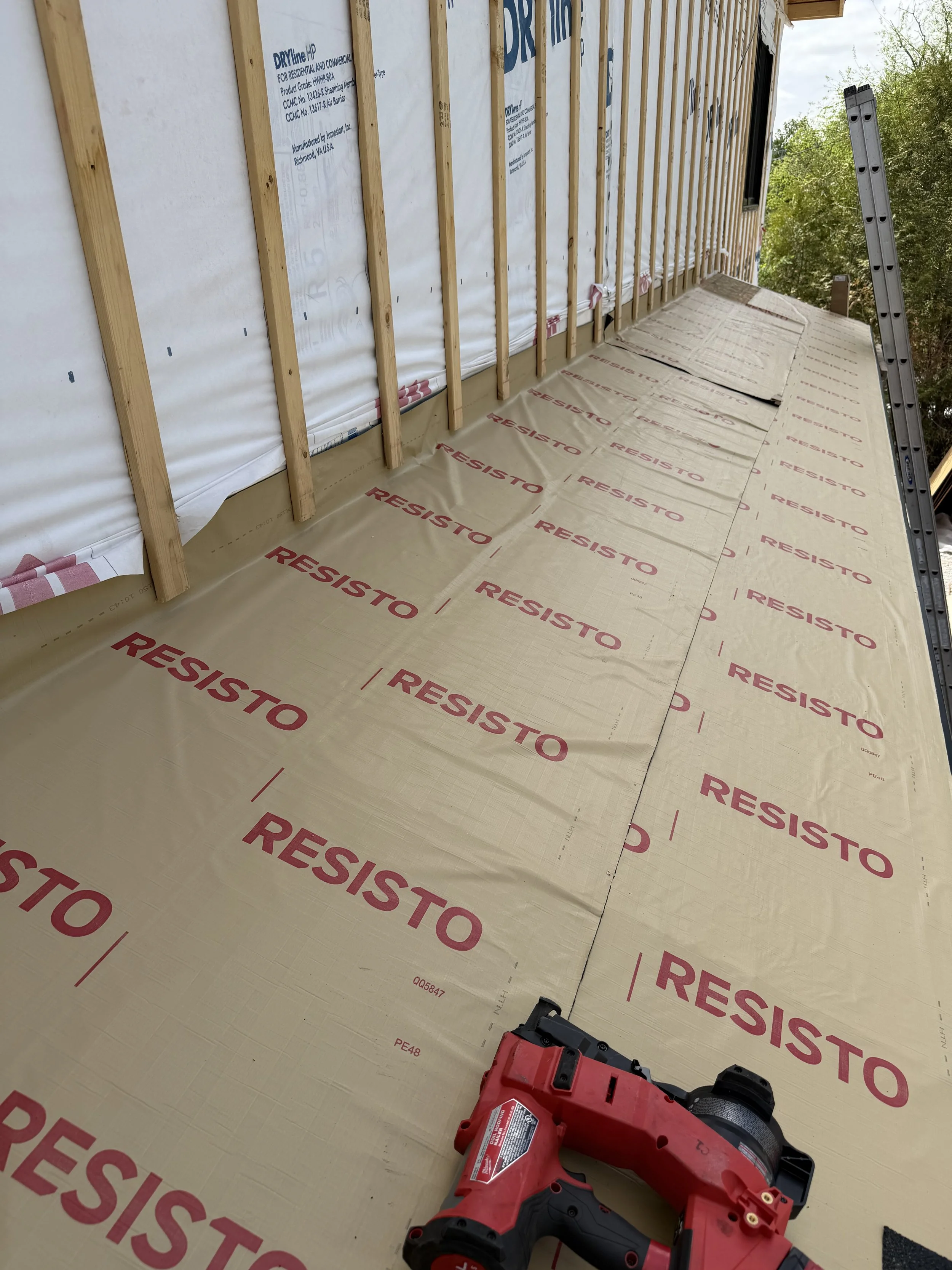 The image shows the interior of a building under construction with exposed wooden framing, a white weather-resistant barrier with the word ‘RESISTO’ printed in red, a ladder outdoors, and a red cordless nail gun on the floor.