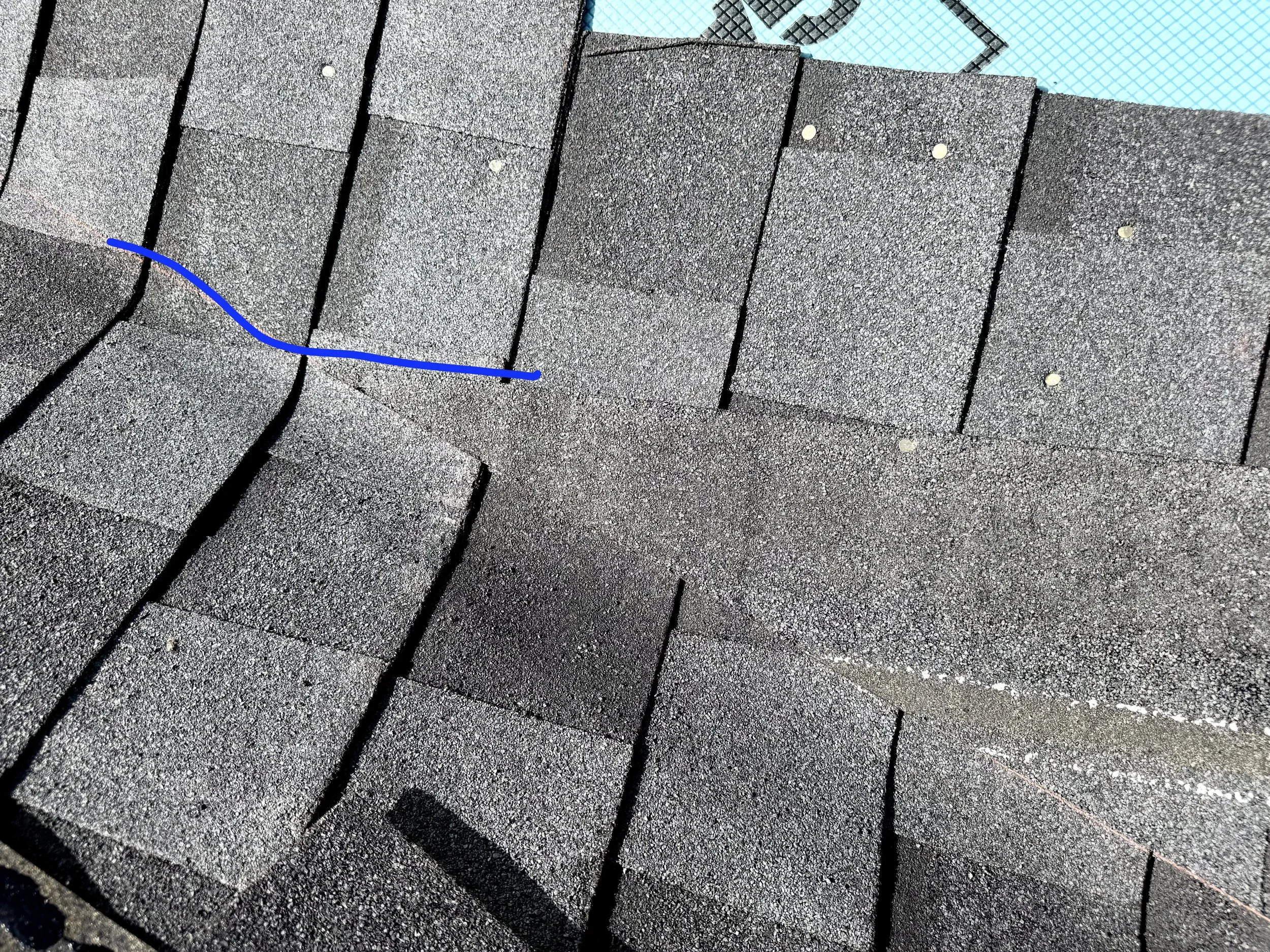 A section of a shingled roof with several small, round, metal nails attached, and part of a swimming pool with blue tiles in the background.