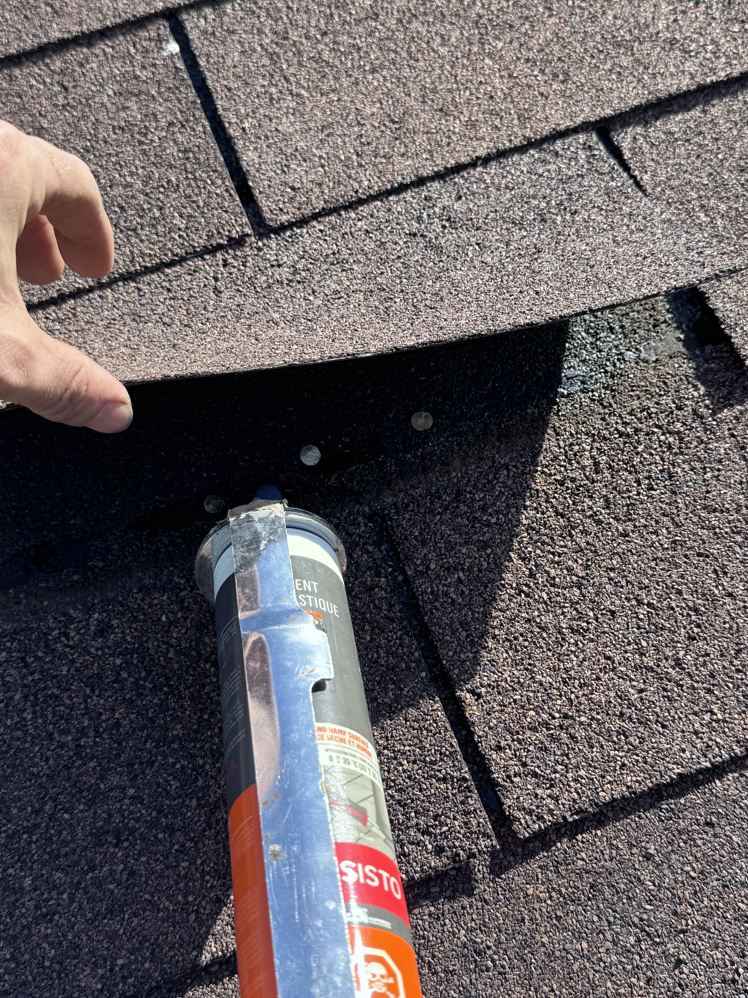 A person's hand with a finger touching a shingle roof. The roof has brown asphalt shingles, and a black trap is visible at the edge of the shingles. Below, there is black asphalt pavement with some loose coins scattered around and a clear plastic bottle with a white cap.
