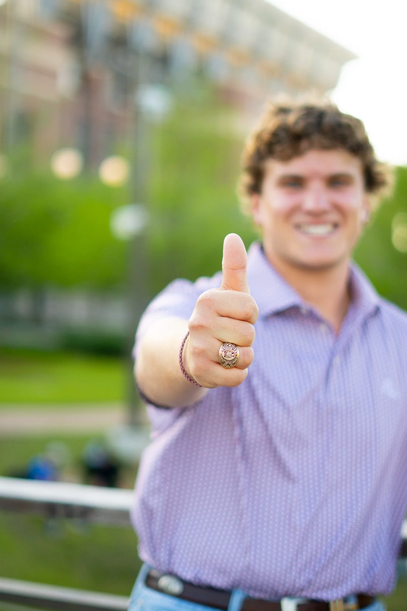 A young man with curly hair smiling and giving a thumbs-up gesture, wearing a checkered shirt and a ring on his finger, outdoors with blurred background of greenery and a building.