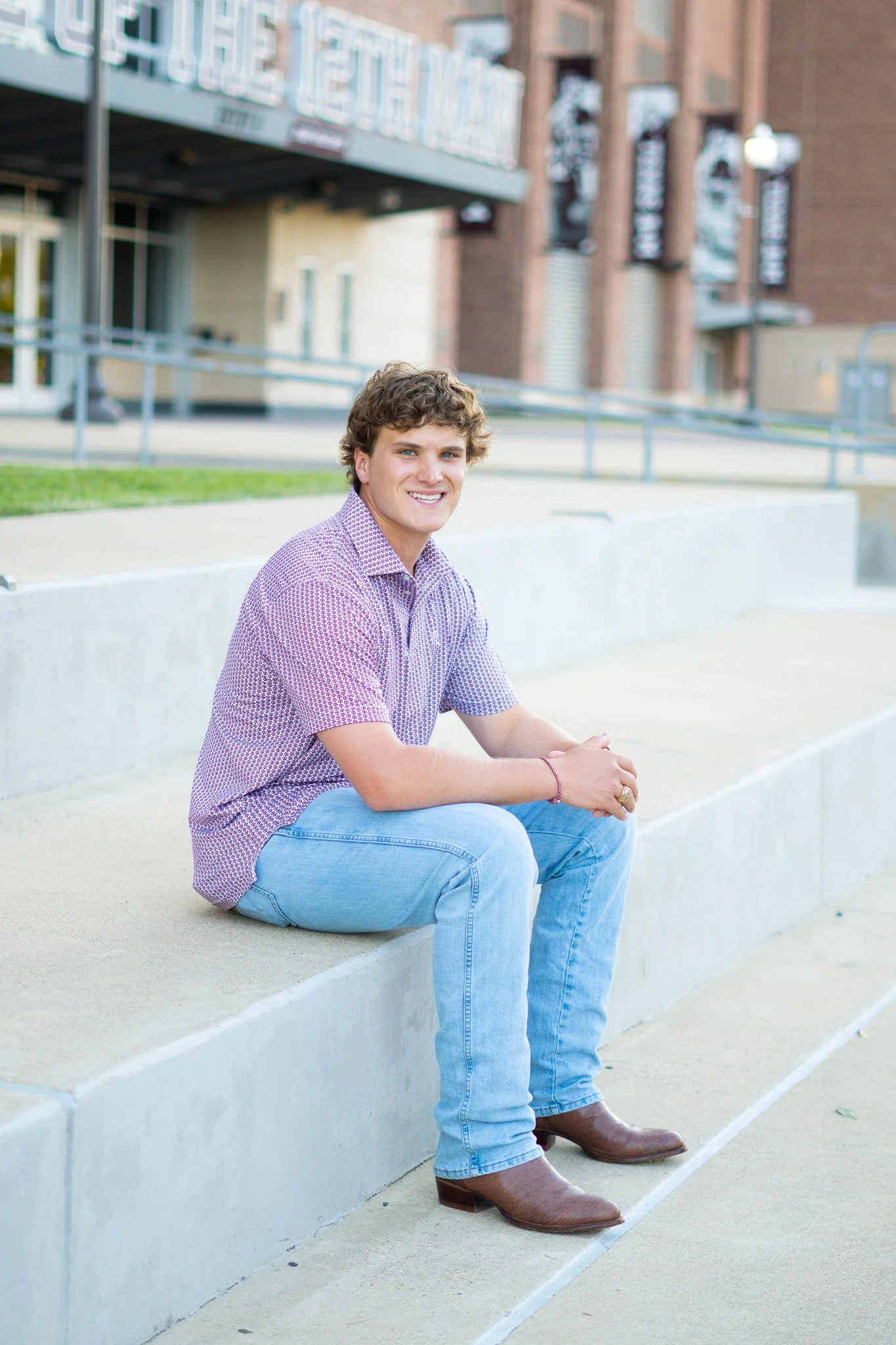 A young man with curly hair sitting on a concrete ledge outside of a building, smiling at the camera. He is wearing a purple patterned shirt, light blue jeans, and brown boots. The building behind him has a modern brick facade with signs and tags, an