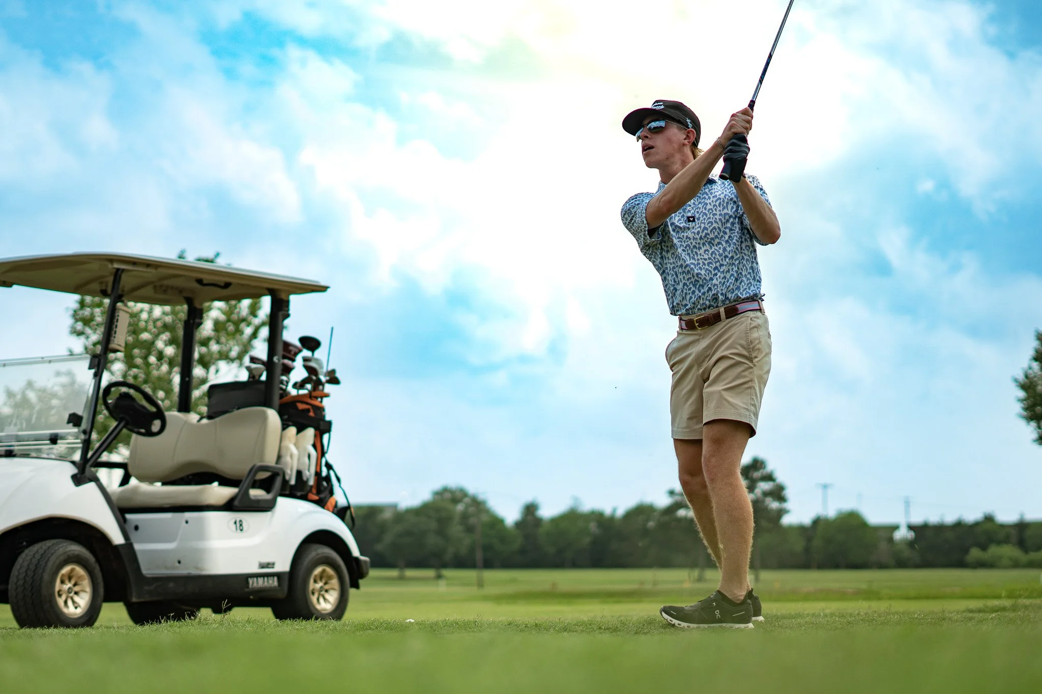 A person in a patterned shirt, beige shorts, and black shoes swinging a golf club on a golf course, with a golf cart loaded with clubs nearby, under a partly cloudy sky.