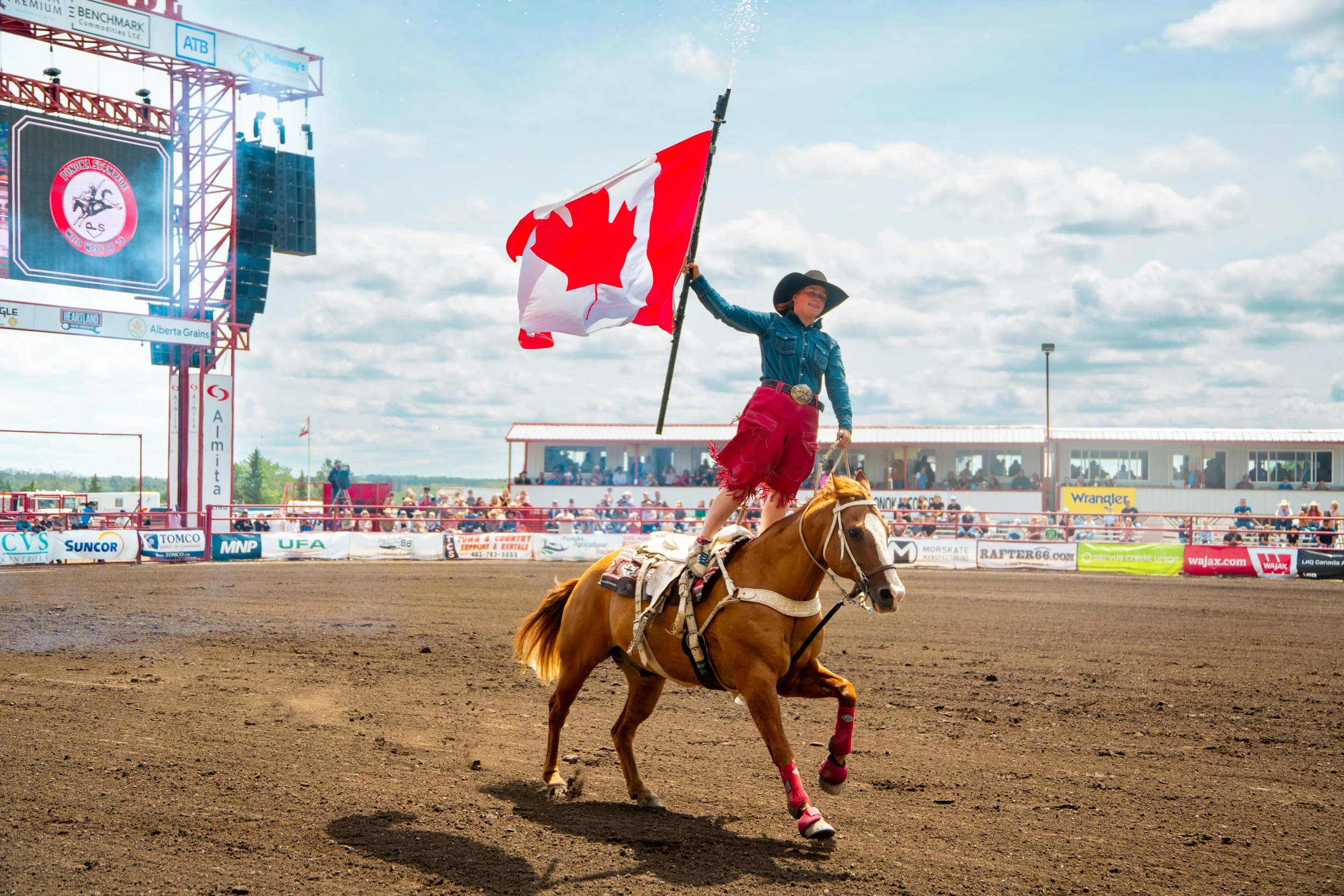 A cowboy is riding a horse in a rodeo arena, holding a Canadian flag and wearing a cowboy hat and jeans, with a crowd watching in the background.