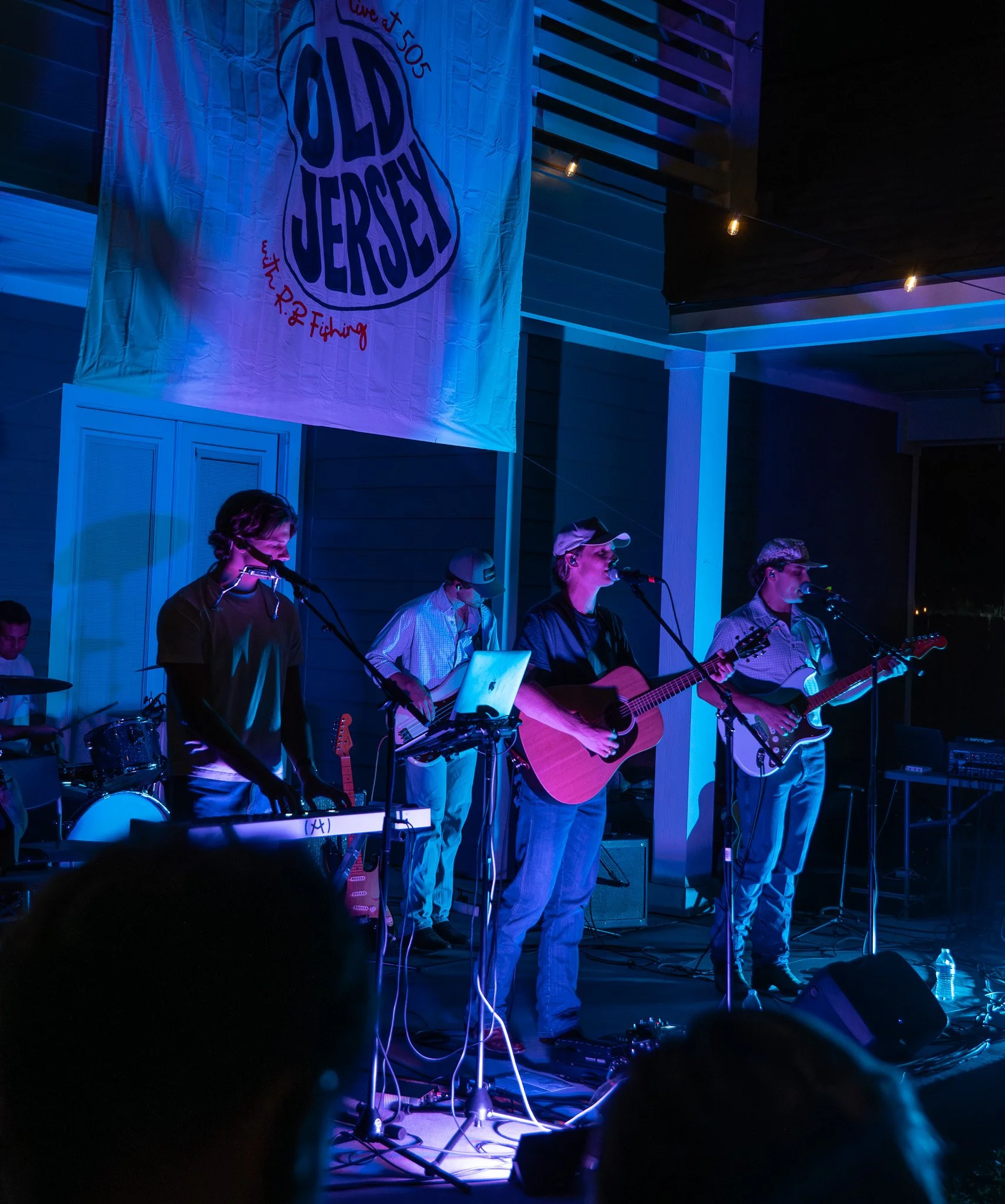 A band of five musicians performing on an outdoor stage at night. The stage is decorated with a large banner that reads 'Old Jersey' and features a fishing-related graphic. Musicians are playing guitar, keyboard, drums, and singing into microphones. 