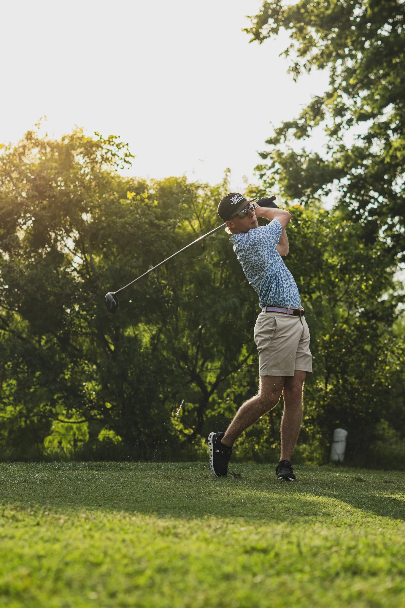 A woman in a blue patterned shirt and beige shorts is playing golf on a lush green course. She is holding a golf club, swinging it, and is wearing sunglasses and a black cap. The background features tall trees and bright sunlight filtering through th