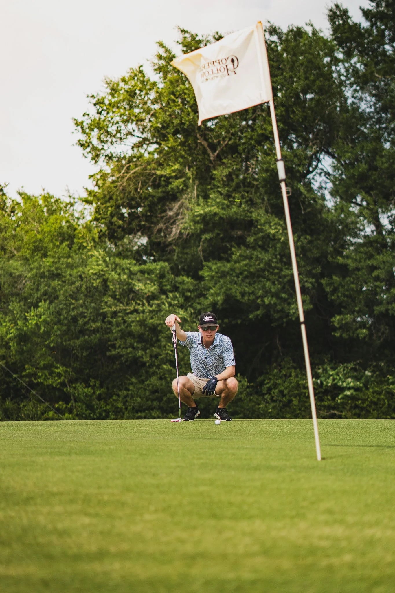 A person in casual clothing crouches on a golf course near the hole, holding a golf club and preparing to putt the golf ball. There is a flagstick with a flag nearby, and lush green trees in the background.