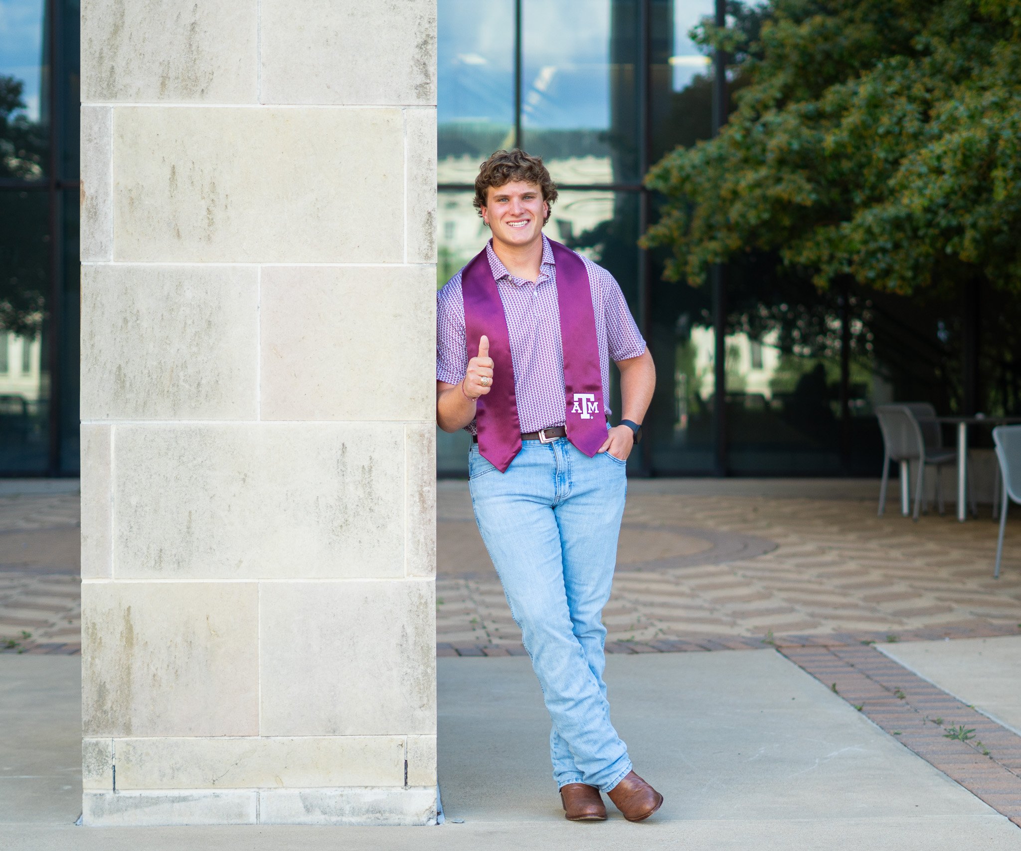 A young man with curly hair smiling, wearing a purple graduation stole with 'TAMU' on it, standing outside next to a stone wall with modern glass building and trees in the background.