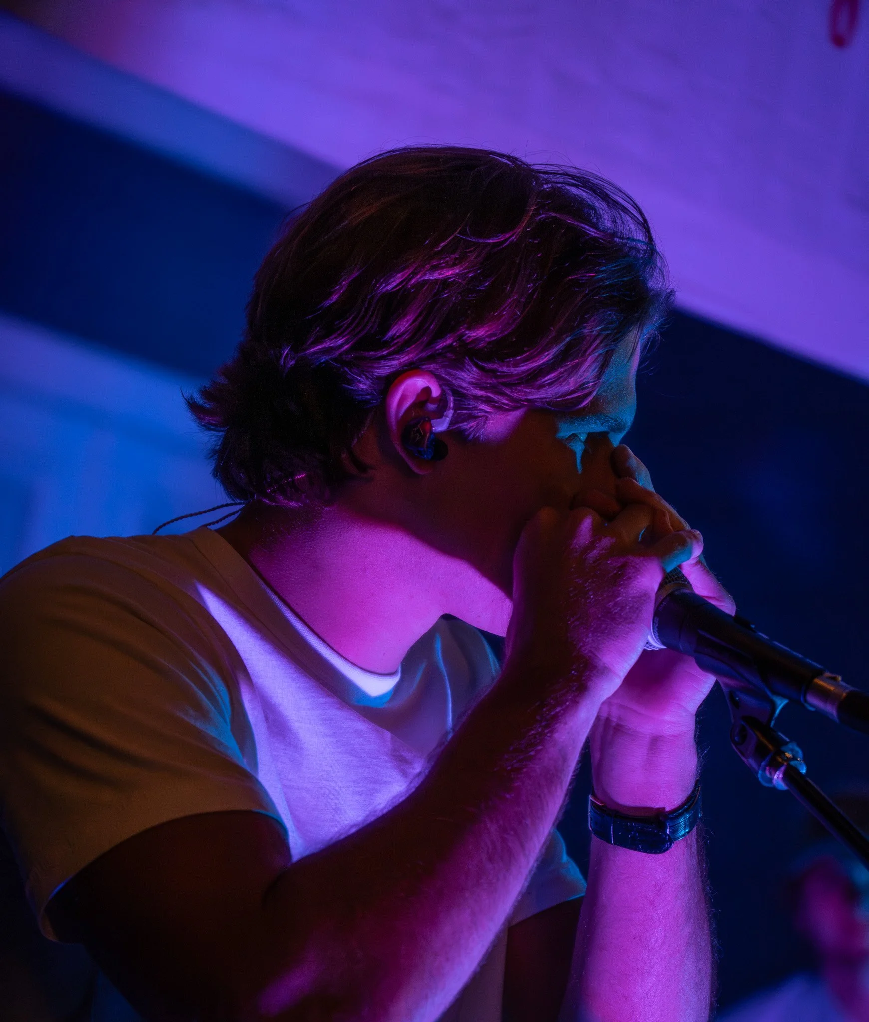 A person with multicolored hair, wearing a white shirt and a brown top, is holding a microphone and appears to be singing or speaking, illuminated by purple and blue lighting.