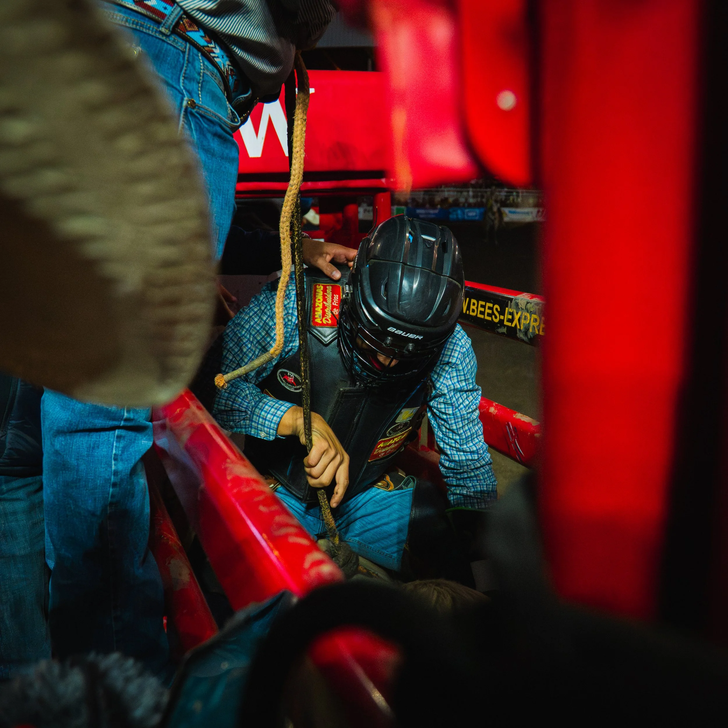 Close-up of a person wearing a black helmet and a plaid shirt, near red objects, possibly on a roller coaster or amusement park ride, with one person holding a strap or rope.