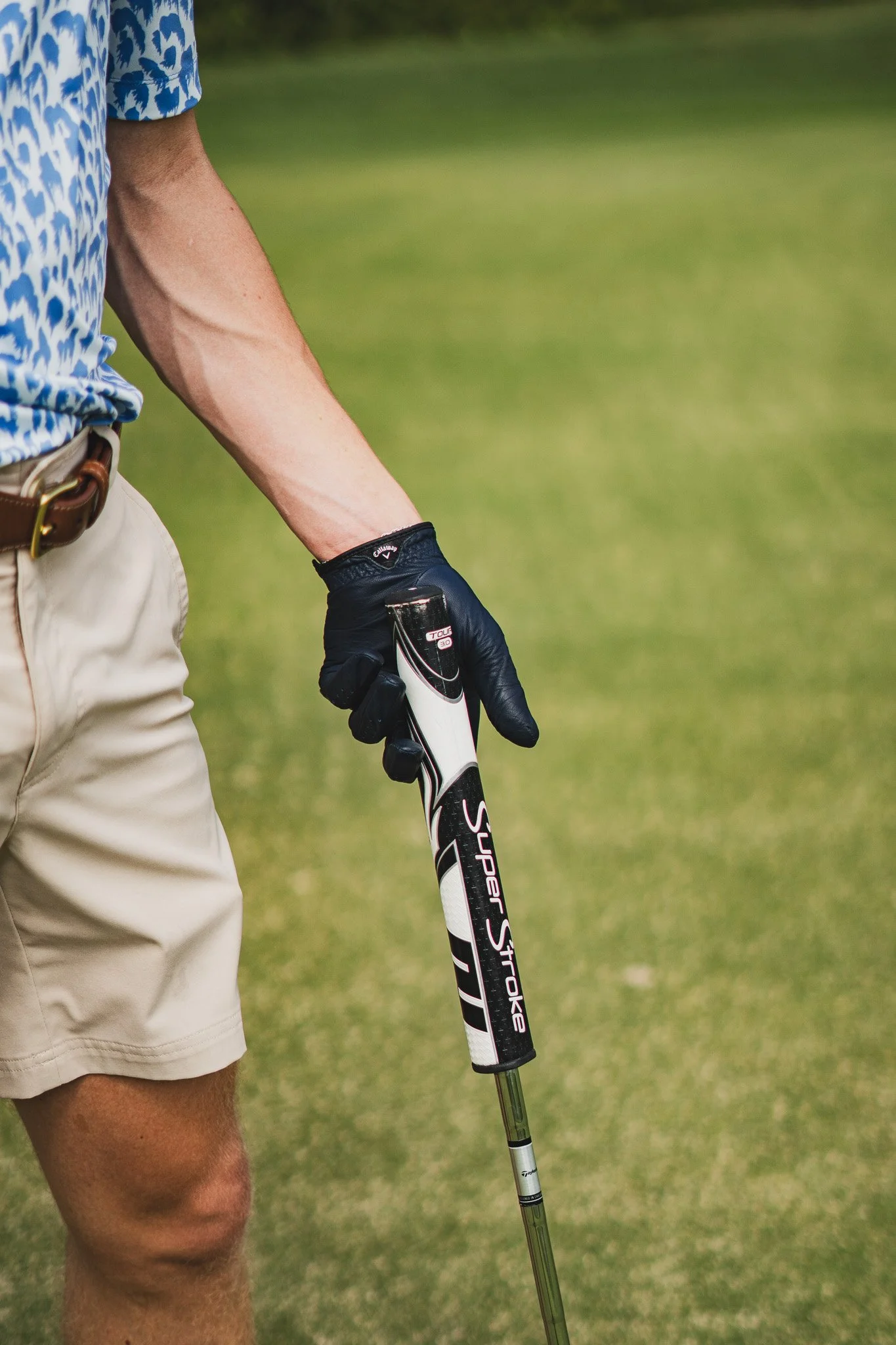 Part of a man wearing a blue patterned shirt and beige shorts, holding a golf club with a black glove, standing on a grassy area.