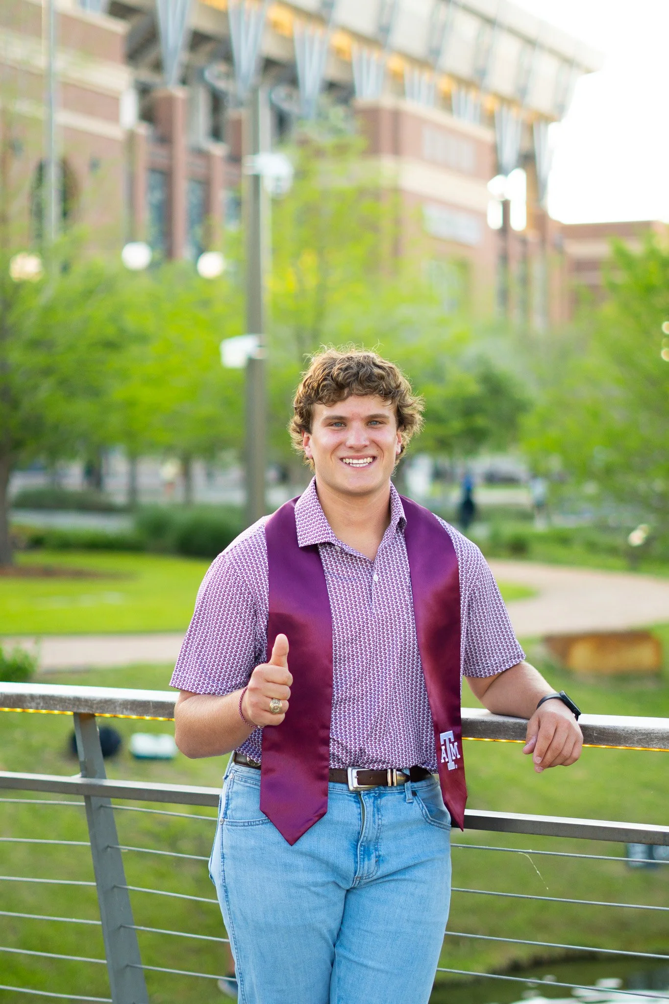 Young man with curly hair smiling and giving a thumbs up, standing outdoors near a metal railing with a university building in the background. He's wearing a purple and white patterned shirt, blue jeans, and a maroon sash with an 'A&M' logo.
