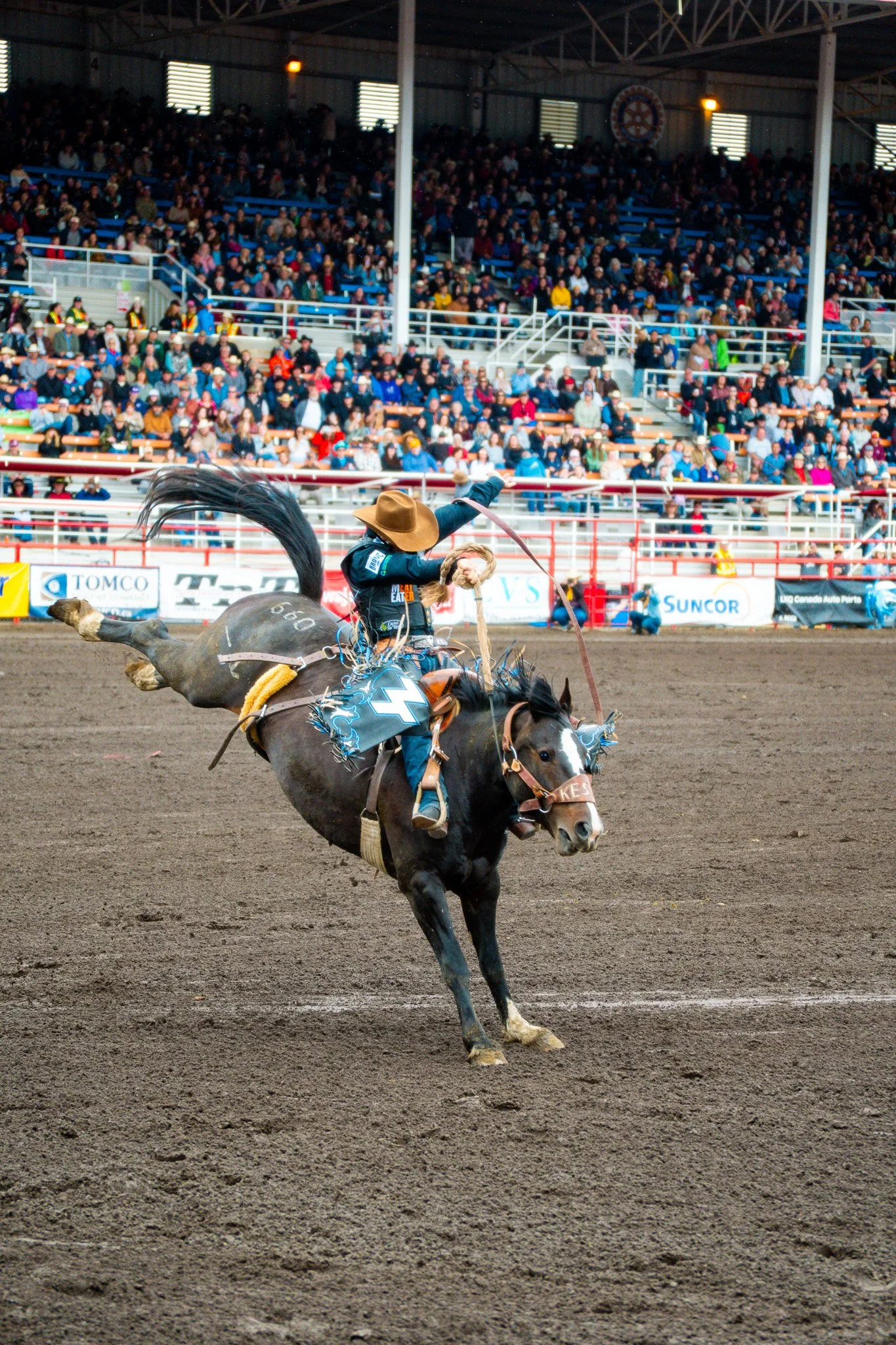 A rodeo cowboy in a hat riding a bucking horse inside an arena filled with spectators.