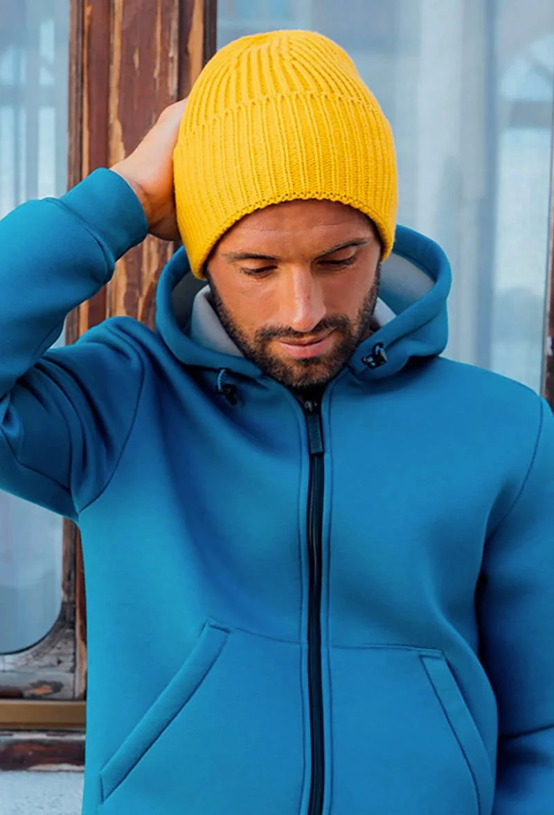 Man wearing a blue jacket and yellow knit beanie, touching the back of his head, standing near a wooden window.