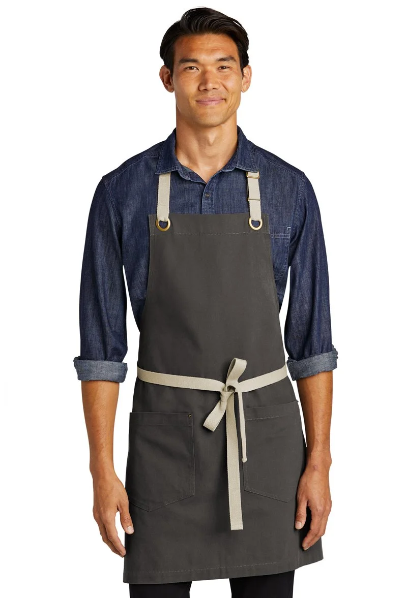 A young man wearing a denim shirt and an apron with a belt, standing against a white background.