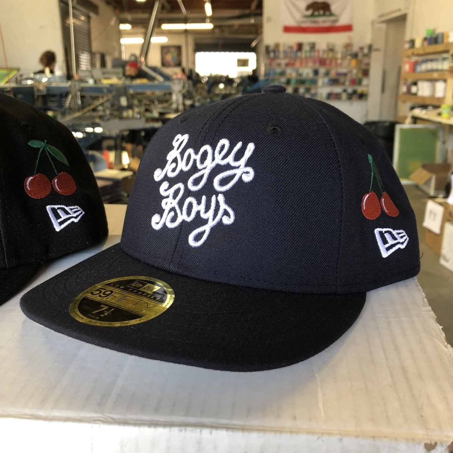 Black snapback hat with embroidered white text 'Joggy Boyz' and cherry graphic on both sides, displayed on a white surface in a store.