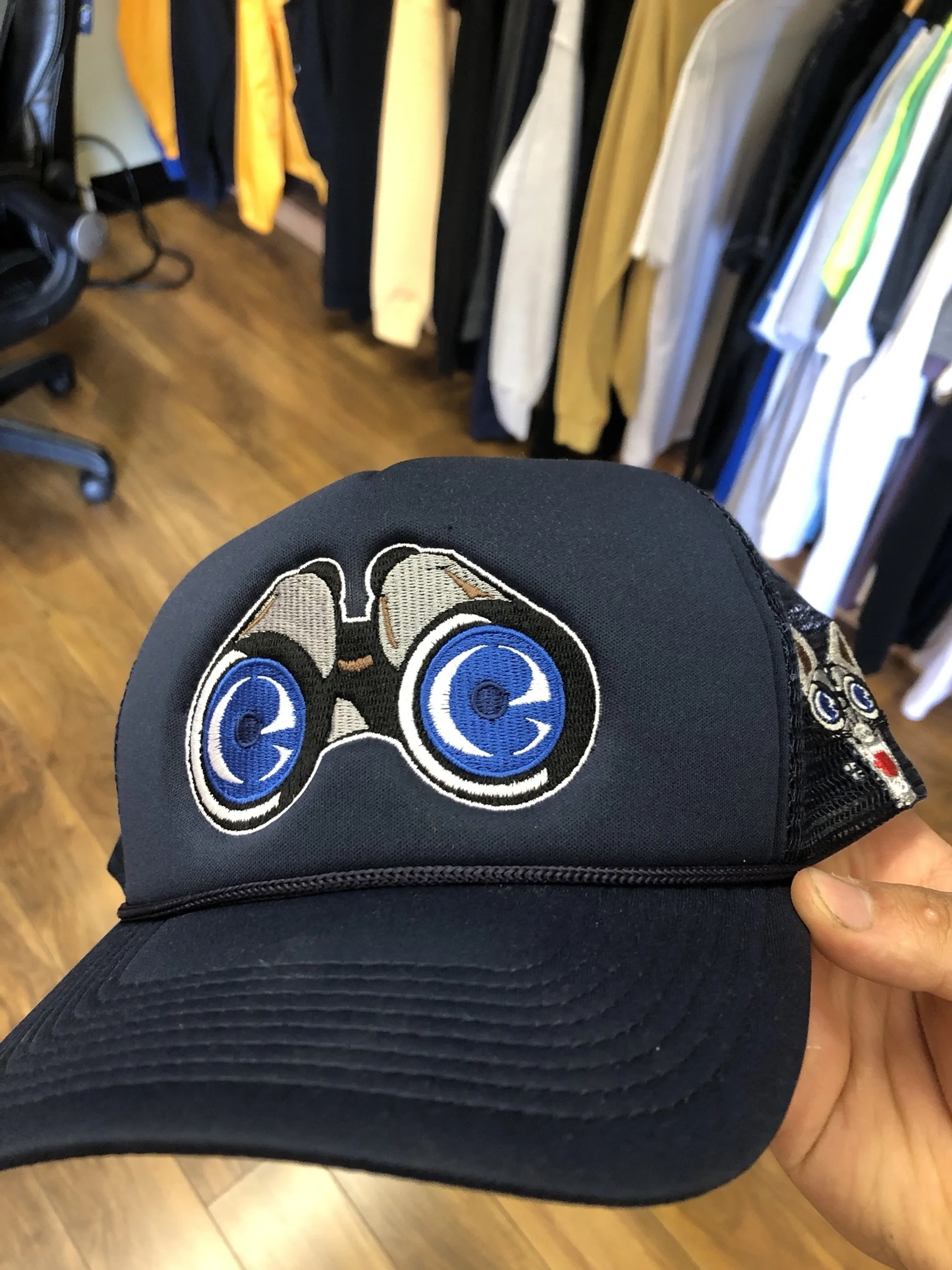 A black baseball cap with a cartoonish embroidered face of binoculars with big blue eyes on the front. The background shows a clothing store with hanging clothes and a wooden floor.
