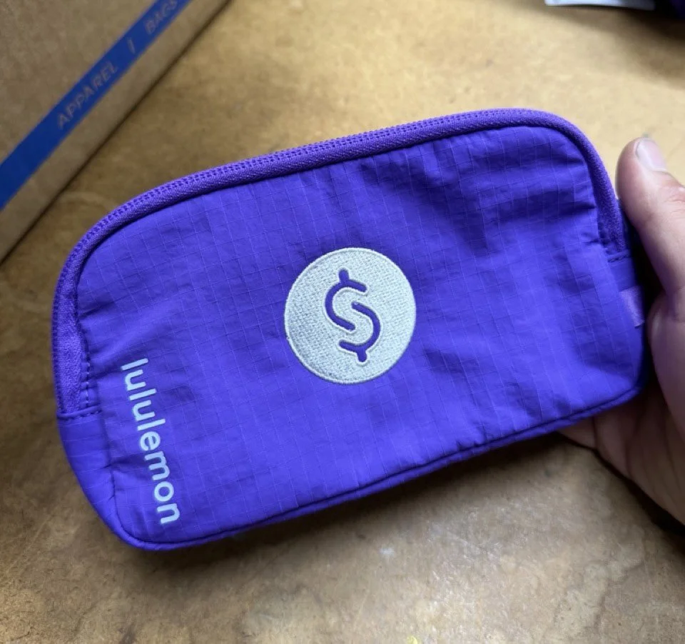 Purple wallet with a dollar sign emblem on the front, held in a person's hand. The wallet is on a wooden surface.
